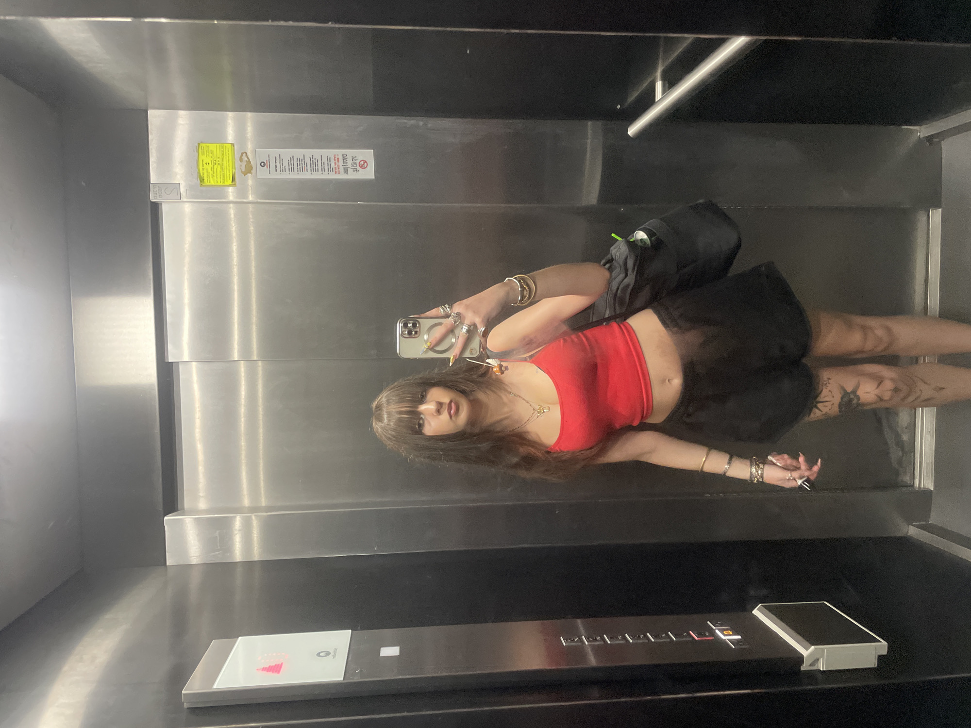 In an elevator in Raanana, Israel, a young woman with long brown hair and bangs stands facing a mirror, taking a selfie. She is wearing a red tank top and black shorts. She has several rings on her fingers and bracelets on her wrists. Her legs have tattoos. She is holding a smartphone in her right hand and has a black bag slung over her left shoulder. The elevator walls are made of brushed stainless steel, and there are buttons and a digital display on the left side of the elevator. A yellow sticker is affixed to the wall on the right side of the elevator.