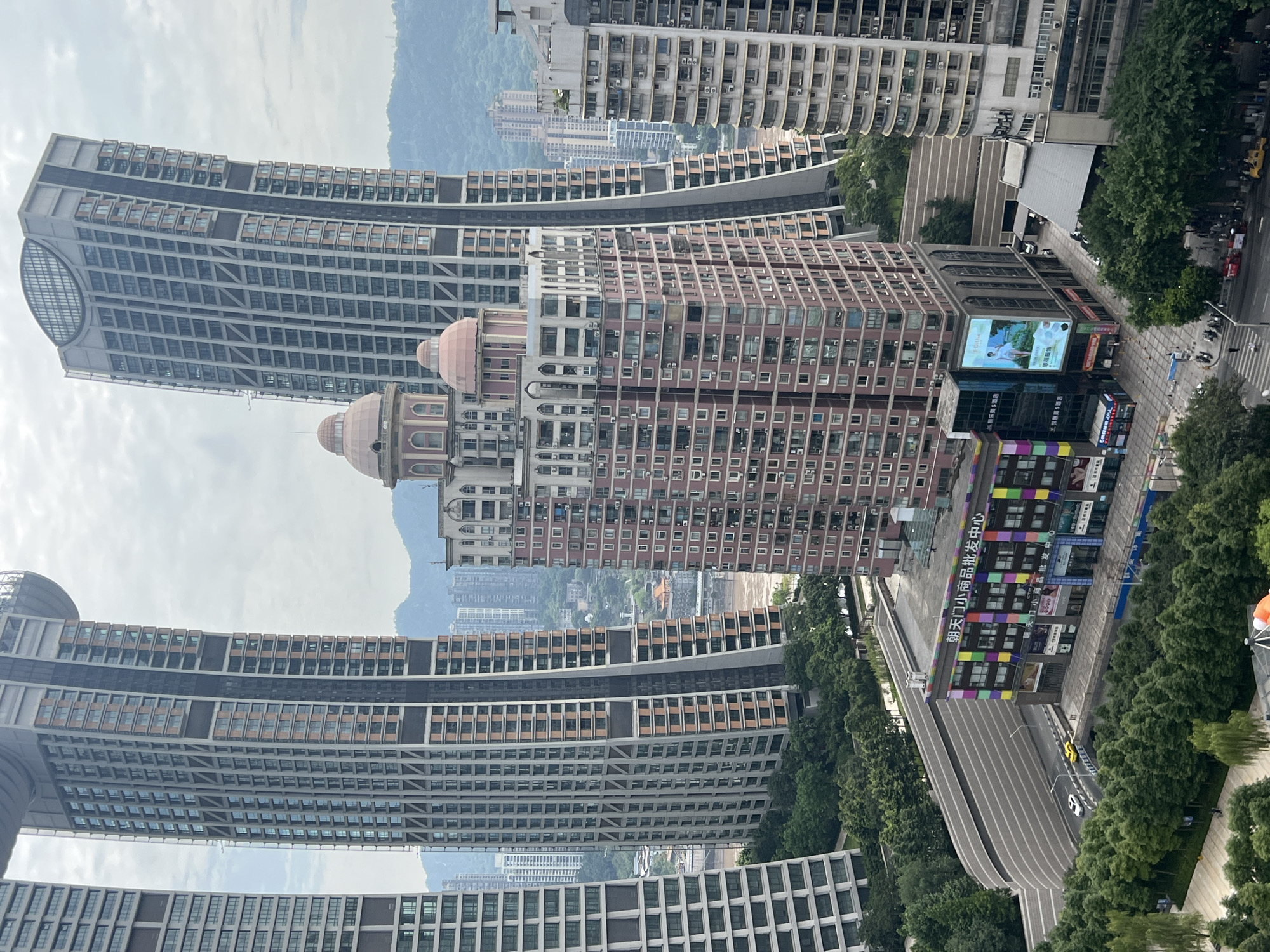 The image captures a dense urban landscape with towering skyscrapers and a prominent building in the foreground. The skyscrapers feature a mix of architectural styles, with some having a sleek, modern design characterized by glass facades and geometric patterns, while others appear more ornate with arched windows and decorative elements. One particularly striking skyscraper on the right is tilted at an angle, giving it a dynamic and unusual appearance.
In the center of the image, a pink-toned building with classical architectural features stands out, topped with two dome-like structures. The lower floors of this building are adorned with colorful, mosaic-like panels and large advertisements, suggesting it may be a commercial establishment or entertainment venue. A prominent sign on this building reads "朝天门小商品批发中心" in Chinese characters, which translates to "Chaotianmen Small Commodities Wholesale Center."
Below the central building, a road curves around a landscaped area with dense green trees. Several cars are visible on the road, including a white car and a yellow car. The background reveals a hazy view of more buildings and rolling hills, indicative of a city nestled within a mountainous region. The sky is overcast, creating a diffused light across the scene. The location is identified as Yuzhong District, China.