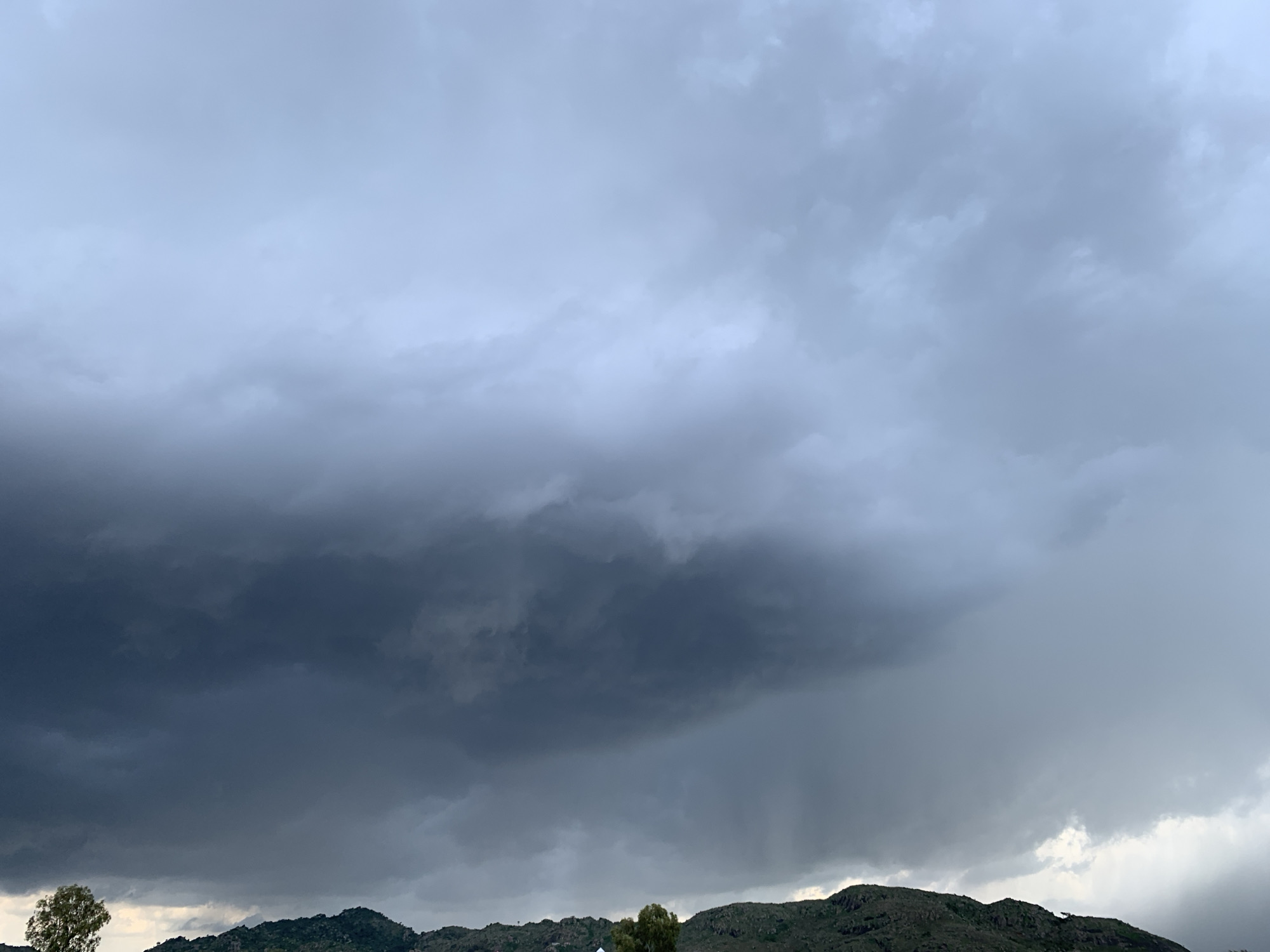 The image captures a dramatic sky over a hilly landscape. The dominant feature is the heavily clouded sky, with dark, ominous storm clouds gathering on the left, gradually transitioning to lighter, more diffuse clouds on the right. These clouds suggest an impending or ongoing rainfall. The landscape below consists of rolling hills covered in vegetation, with a few trees visible in the foreground. The overall lighting is dim and diffused, consistent with overcast weather. The scene evokes a sense of anticipation or the aftermath of a storm. No people, objects, or text are discernible in the image. The location context suggests Kuru Karama, Nigeria.