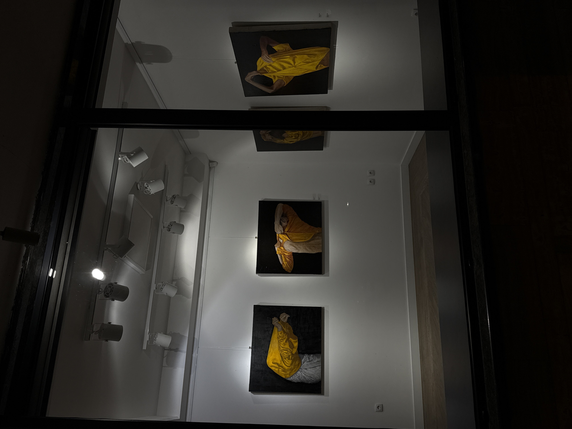 The image shows an interior scene, likely a gallery or exhibition space in Belgrade, Serbia, based on the architectural style and the presentation of artwork. The view is partially obstructed by a dark frame, possibly a window or doorway.
On the left, a wall features a series of adjustable spotlights. The wall facing forward displays four square canvases, each featuring a portrait of a person draped in golden-yellow fabric. The figures and fabric are rendered in high contrast against a dark background, creating a dramatic effect.
The right side of the image is dark, suggesting a neighboring room or a darkened part of the gallery. The overall lighting is subdued, with the focus directed towards illuminating the artwork.