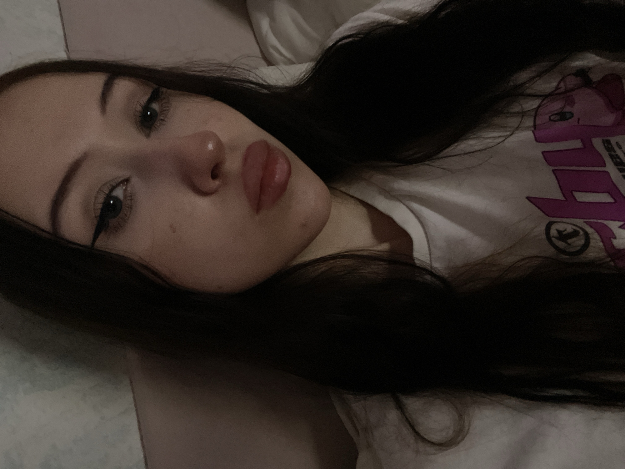 A close-up, low-light, indoor selfie shows a young woman with long, dark hair. She is wearing a white t-shirt with a pink graphic on it, featuring a cartoon character that resembles Kirby. The woman has prominent eyeliner and her lips are slightly parted. The background is blurred, with hints of a textured wall and a soft, possibly fabric, surface. The image's setting is not specified as being in Votkinsk, Russia, and no other geographical or contextual information is provided.