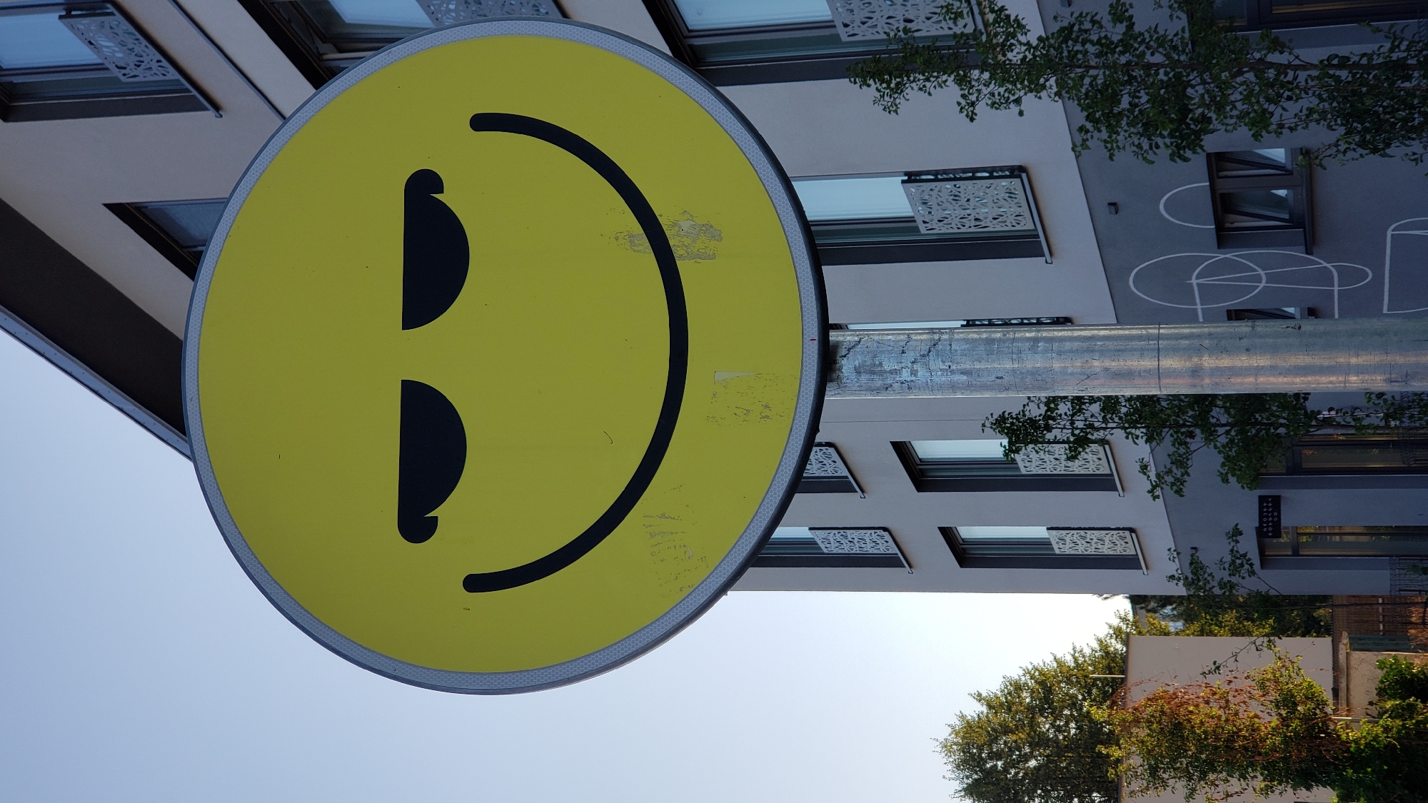 A yellow, oval-shaped sign with a black smiley face is mounted on a metal pole in front of a modern building. The smiley face has closed, angled eyes, giving it a mischievous or cool expression. The sign is slightly worn, with some scuff marks visible. The building behind it is light gray with multiple windows and decorative metal railings on some of the balconies. Trees and greenery are present on the left and right sides of the image, suggesting an outdoor urban environment. The sky is clear and blue. This image was taken in Prešov, Slovakia.