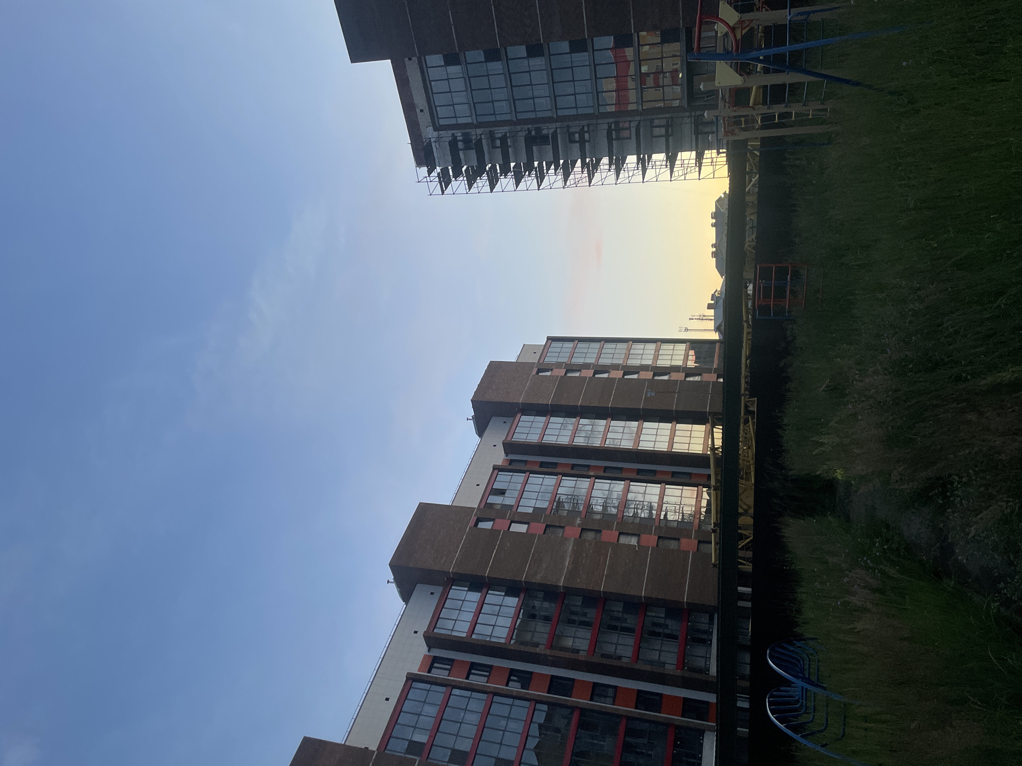 The image captures an outdoor scene with multiple tall buildings under a clear blue sky. The buildings are modern, featuring a repeating pattern of windows with dark frames and some red accents. One building, in the foreground and on the left, is viewed from a low angle, emphasizing its height. Another building, further in the background and on the upper right, appears to be under construction, with scaffolding visible on its exterior. Between the buildings, a playground structure with blue and yellow elements is partially visible, situated on a grassy area. The overall impression is that of an urban or suburban residential area, possibly a newly developed one, given the construction. The lighting suggests it might be late afternoon or early evening, with the sun casting a warm glow in the distance, particularly on the upper right building and the sky. The grass in the foreground is tall and somewhat unkempt. There are no people visible in the frame. No text is discernible. The location context provided is Laishevo, Russia.