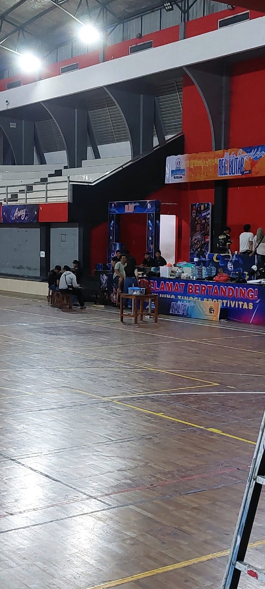 The image depicts an indoor sporting venue, likely a gymnasium, with a wooden floor marked with various lines. In the mid-ground, a group of people are gathered around tables and some are seated on wooden stools. A banner is displayed above them with the text "SELAMAT BERTANDING!" which translates to "Good luck competing!". Another banner to the right reads "REG. BLITAR". In the background, there are elevated seating areas and architectural elements of the building's structure, including lighting fixtures. The scene appears to be set up for an event or competition.