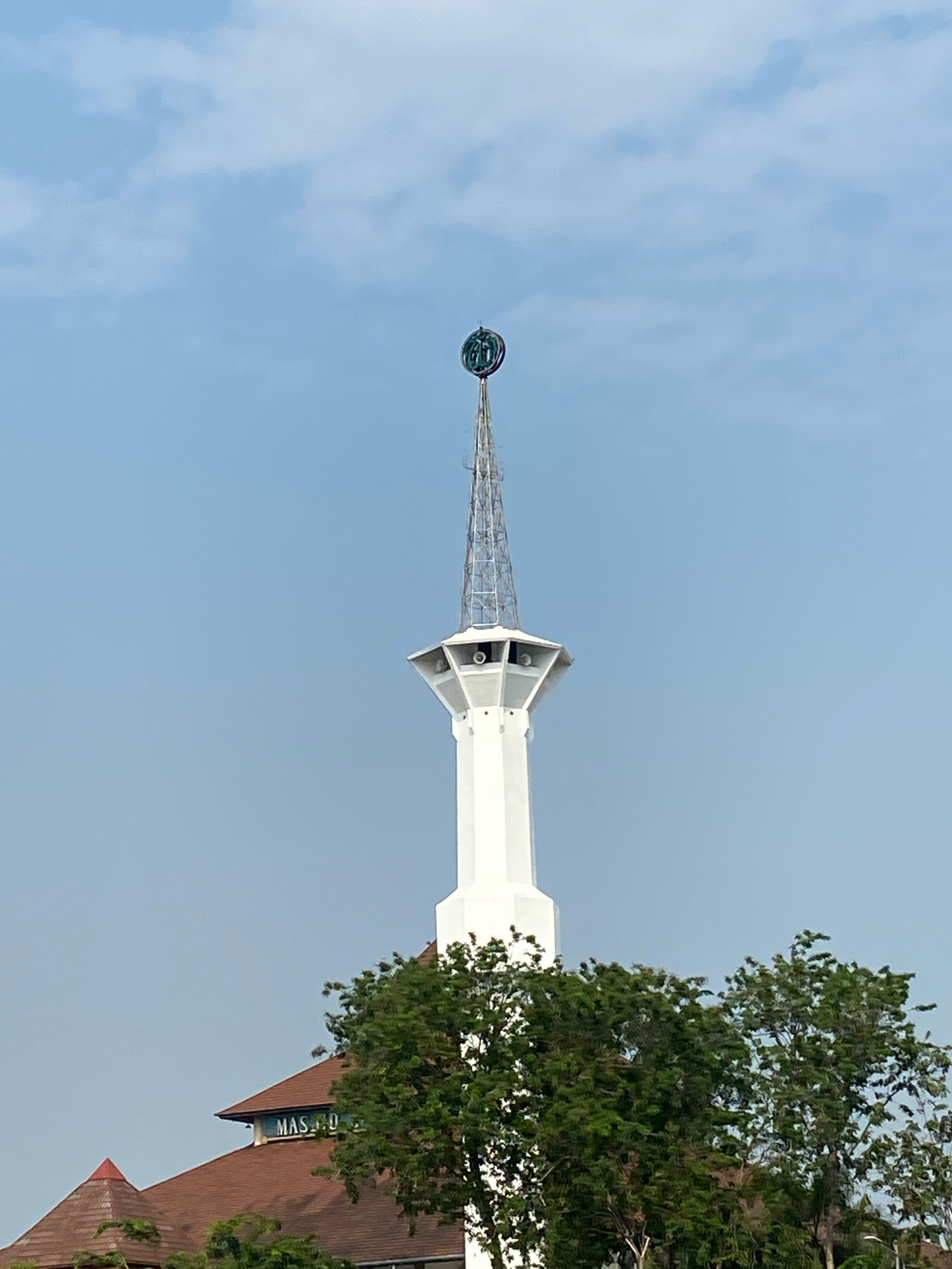 The image shows the minaret of the Masjid Agung Sidoarjo, a prominent mosque in Sidoarjo, Indonesia. The minaret is tall and white, with a lattice-like structure at the top supporting a decorative element. The sky is clear and blue, with a few wispy clouds. In the foreground, the roof of the mosque is visible, along with lush green trees. The overall scene is serene and captures the architectural beauty of the mosque.