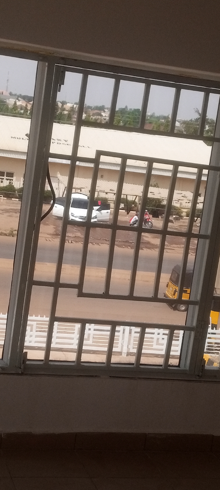 The image depicts a view from a window with a metal grate, overlooking a street scene in what is likely Sabon Tasha, Nigeria.
Objects: A white building, a white car with the driver-side doors open, a motorcycle with two people, an auto rickshaw (yellow and black), a white fence, a window with metal grate.
Environment: A street surface appears to be a combination of paved and unpaved areas, with possible dirt or debris present. There is a mix of buildings and vegetation. The sky is overcast.