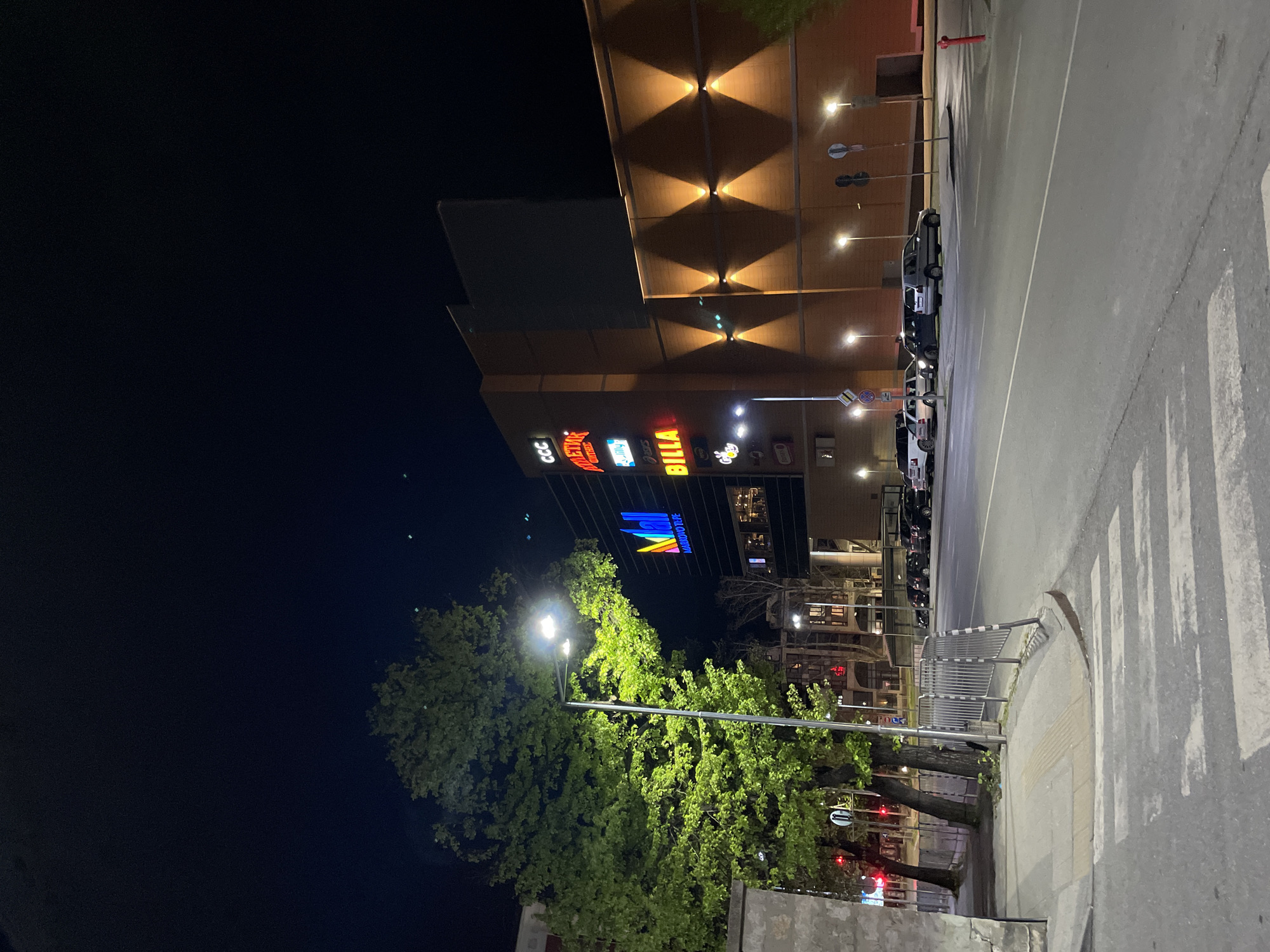 Nighttime scene in Plovdiv, Bulgaria, featuring a multi-story building with illuminated storefronts including "CCC," "Deichmann Outlet," "Billa," and "Marriott." The building facade is decorated with diamond-patterned lights. The street beside the building is well-lit with streetlights and features parked cars along the side. A pedestrian crossing is visible in the foreground. Trees line the street, partially obscuring lower buildings in the background. The sky is dark.