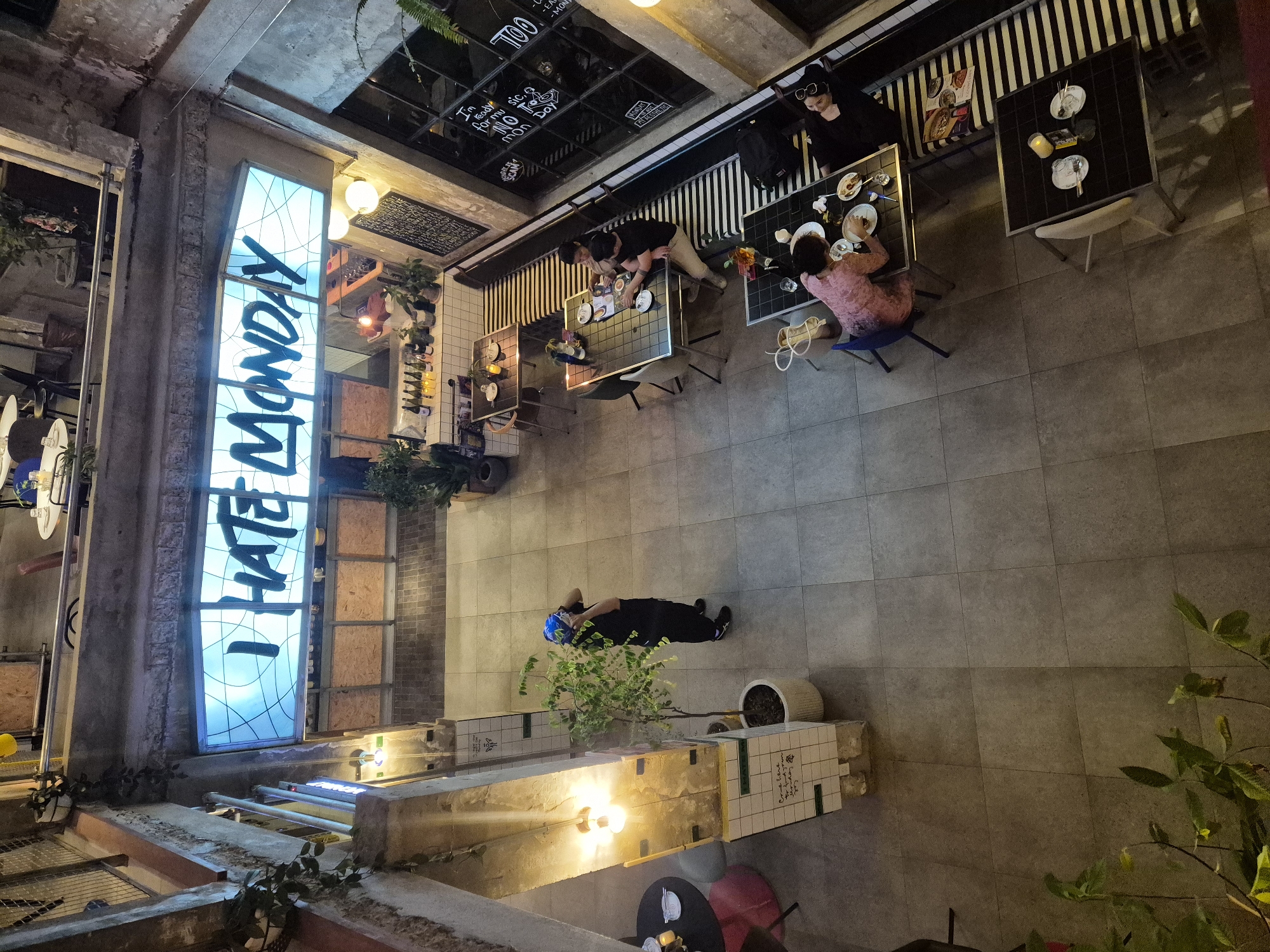 This is an overhead, wide-angle shot of an indoor cafe in Da Nang, Vietnam, captured at night. The cafe features an industrial-chic aesthetic with exposed concrete walls and pillars. A prominent, illuminated sign above reads "I HATE MONDAY" in large, blue, sans-serif font.
In the foreground, a person wearing a blue helmet and a dark outfit stands near a potted plant, appearing to take a photograph with their phone. Throughout the cafe, several tables are occupied by patrons.
On the left side, a round table with two chairs and a pink stool is set with plates and a candle. Further back, another table is occupied by a man and a woman sitting opposite each other, looking at a menu.
To the right, a long bench with black and white stripes accommodates two women. One woman, wearing a black top, is reclined and appears to be resting her head on her hand. The other, in a pink dress, is seated and looking down at her meal.
Another smaller, dark-colored table with two chairs is visible in the bottom right corner, set with plates and a candle.
The floor is tiled with large, gray square tiles. Plants are scattered throughout the space, adding a touch of greenery. Bottles of wine are arranged on shelves behind the bar area, and some decorative chalkboards are also visible. The overall lighting is dim, creating a moody atmosphere, with spotlights and the large neon sign providing the primary illumination.