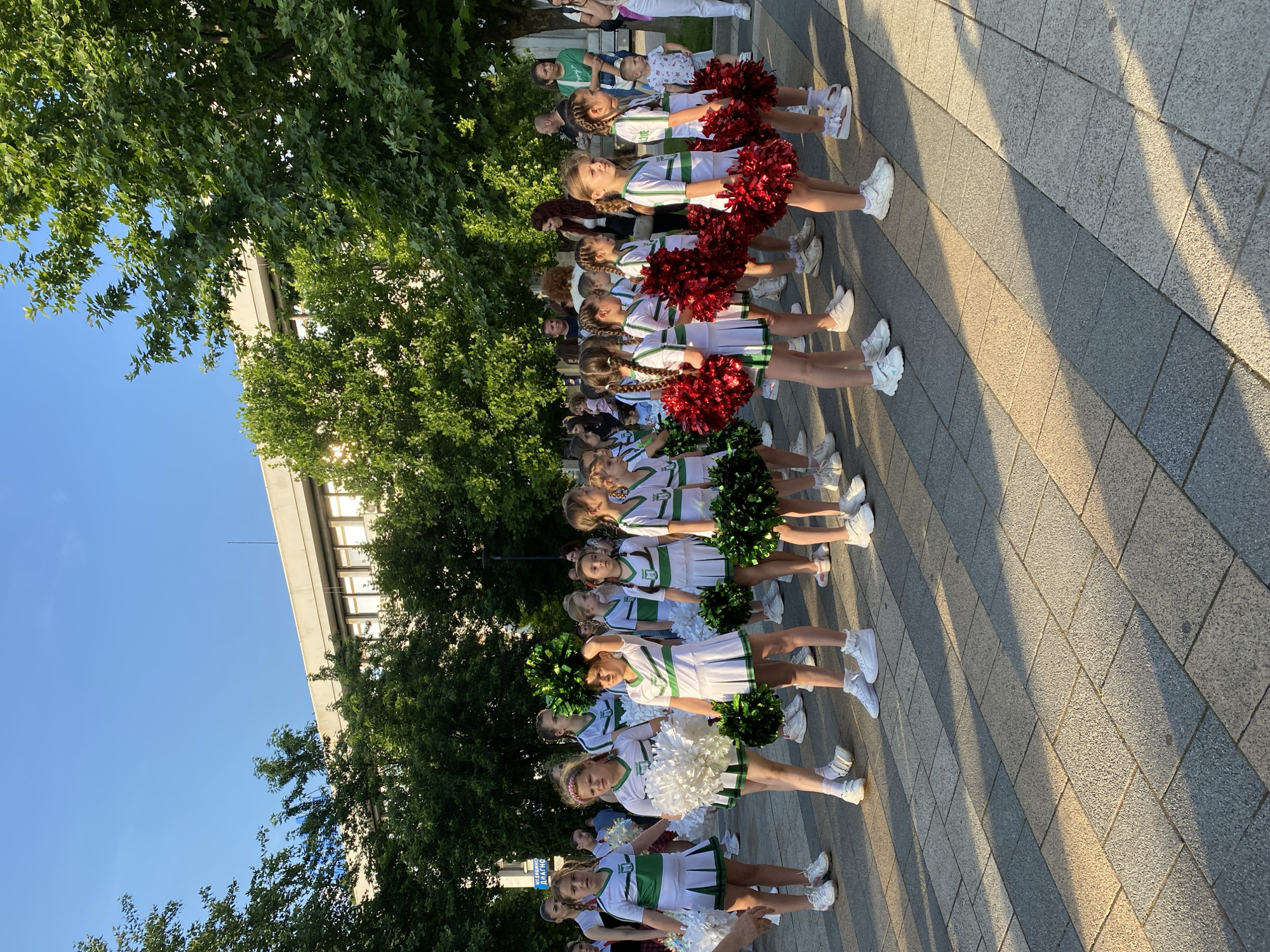 This image captures an outdoor scene in Pleven, Bulgaria, likely a public plaza or pedestrian area, under bright late afternoon or early evening sunlight.
The primary subjects are a group of approximately two dozen young girls, appearing to be cheerleaders, ranging in age from roughly 7 to 12 years old. They are dressed in identical white short-sleeved tops featuring a horizontal green stripe across the chest, a green V-neck trim, and a green shield-shaped emblem on the left chest. They wear white pleated skirts with a green band along the hem, and white athletic shoes with white socks. The girls are arranged in several rows, generally facing forward and slightly to the right of the camera's perspective, standing in a formation.
The girls hold various colored pom-poms:
* The group on the far left holds white pom-poms.
* The central group holds dark green, bushy pom-poms, some raised above their heads.
* The group on the right holds vibrant metallic red pom-poms.
The ground is paved with rectangular interlocking pavers laid in alternating light grey and darker grey strips, running perpendicular to the line of cheerleaders. Long, distinct shadows are cast across the pavement by the cheerleaders and trees, indicating a low sun angle from the left side of the frame.
In the mid-ground and background, several mature deciduous trees with dense green foliage are visible, partially obscuring a large, multi-story modernist building with numerous rectangular windows and a flat roof. A portion of a blue flag or banner is visible above the trees on the far left. A sign on the left side, behind the cheerleaders, contains Bulgarian text, including "МЕДИЦИНСКО ДИАГНОСТИЧЕН" (Medical Diagnostic).
Various adults and a few younger children are present in the background, some observing the cheerleaders, others walking or standing casually. They are dressed in casual street attire. The overall atmosphere suggests a public gathering or performance. The sky above the building and trees is clear and blue.