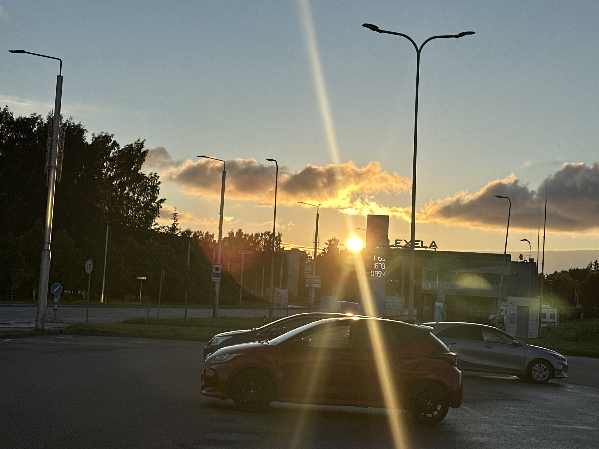 The image captures a sunset scene at a gas station in Tallinn, Estonia. The sky is filled with warm hues of orange and yellow as the sun sets behind a row of trees and a building. Several streetlights are visible, some silhouetted against the sky and others illuminated by the fading sunlight. Two cars are parked at the gas station, one red and one silver, with their headlights on. The gas station itself appears to be part of a larger commercial area, with a sign visible that reads "LEXELA". The overall atmosphere is one of a calm evening in an urban setting.