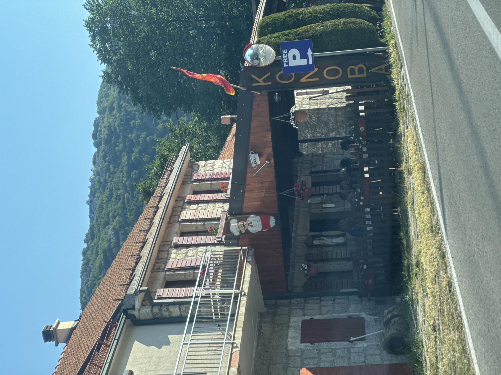 This is a photograph of a building in Njeguši, Montenegro. The building appears to be a restaurant or tavern, with a rustic stone and wood facade. A wooden sign with gold lettering that reads "KONDA" is visible, along with a blue sign indicating "FREE PARKING" with a white arrow pointing right. A red and gold flag is flying from a pole attached to the building. The building has red-tiled roofs and wooden shutters on the windows. There is a balcony with a metal railing on the upper floor. In front of the building, there is an outdoor seating area enclosed by a wooden fence. Several potted plants with red and white flowers are visible on the fence and balconies. A wooden cutout of a chef is mounted on the exterior wall. In the background, a lush green hillside with trees and rocky outcrops can be seen under a clear blue sky. The foreground shows a paved road with a white line marking the edge. The overall scene suggests a daytime, sunny setting in a rural or mountainous area.
