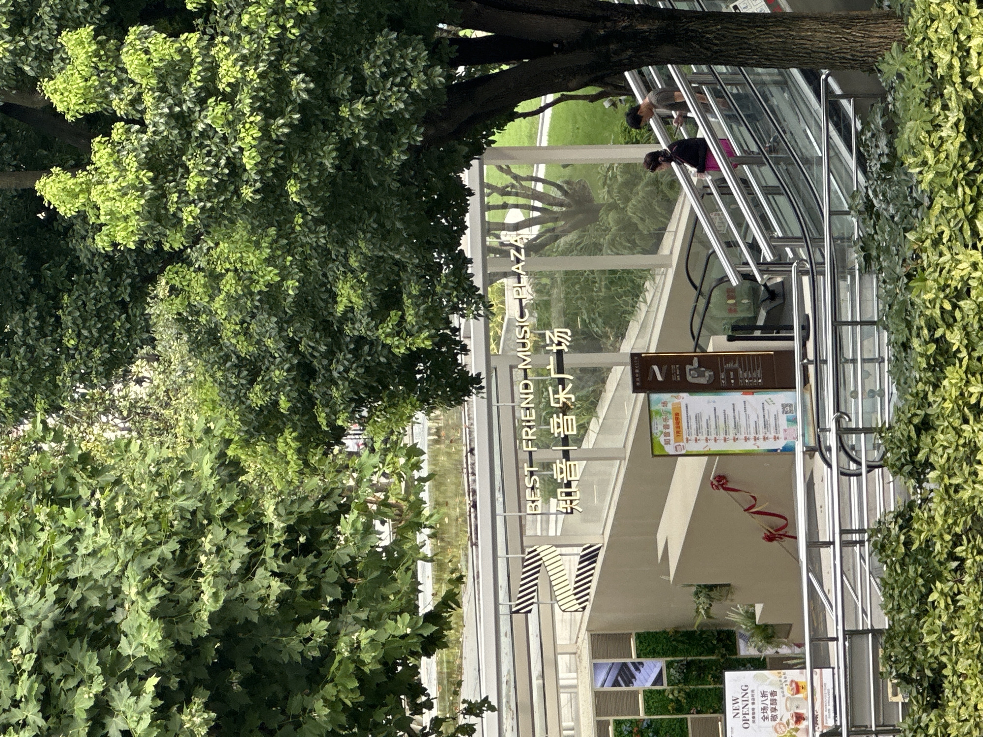 The image depicts the entrance to "BEST FRIEND MUSIC PLAZA" in Shanghai, China. The plaza's name is visible in English and Chinese characters, "知音|音乐广场". An escalator is in operation, with two people, a woman in a pink skirt and a man in a gray t-shirt, ascending. To the left, there's a green wall with a decorative piano keyboard design. A red ribbon decoration is tied to a railing on the stairs to the left of the escalator. A sign with information about a "NEW OPENING" with a discount is also visible. Lush green trees and foliage surround the building, suggesting an outdoor, possibly urban, environment.