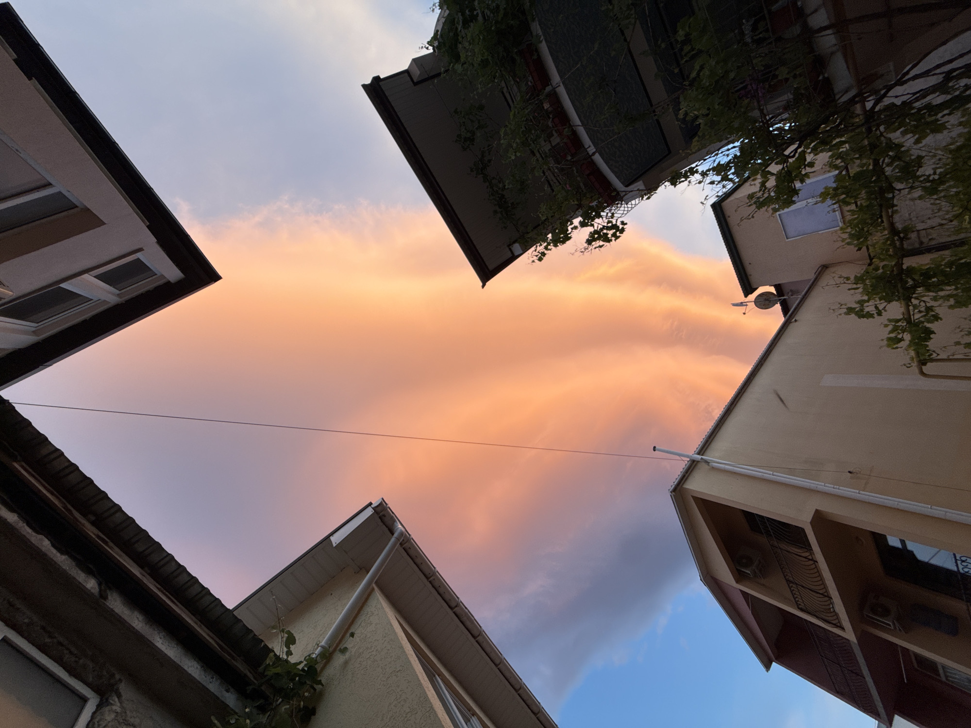 The image captures a low-angle view of a sky, framed by the roofs and upper stories of buildings. The sky is predominantly filled with pink and orange clouds, suggesting either sunrise or sunset. Patches of blue sky are visible in the lower right corner. The buildings are various shades of beige, brown, and grey. Some have foliage growing on them. Power lines stretch across the sky in the center. Air conditioning units are visible on one of the buildings. The location is identified as being in the city of Unknown, Unknown.