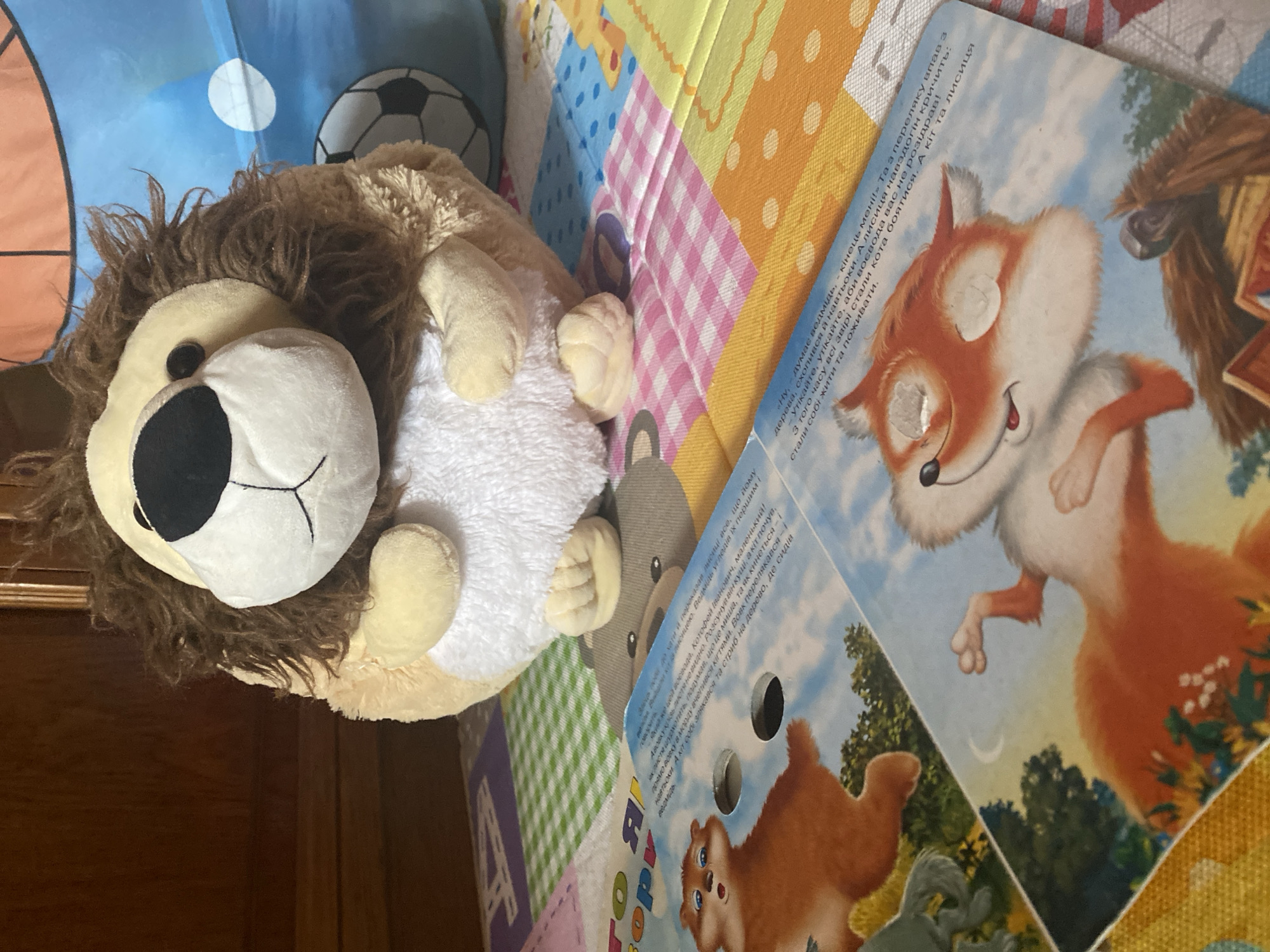 The image features a stuffed lion toy placed on a colorful mat with a book. The mat has a patchwork design with various colors and patterns, including pink and white checks, orange dots, and green squares. The book, positioned on the right side of the image, displays illustrations of cartoon animals, including a fox and a bear, and has text in Cyrillic script. The book is open, revealing its pages. The stuffed lion is positioned on the left, with its face prominently visible. It has a brown mane, a white face with a black nose and eyes, and beige paws. The scene appears to be indoors, possibly a child's play area, given the presence of the toy and the colorful mat. The lighting suggests it might be daytime. The overall impression is one of a playful and cozy environment. The text on the book appears to be a children's story.