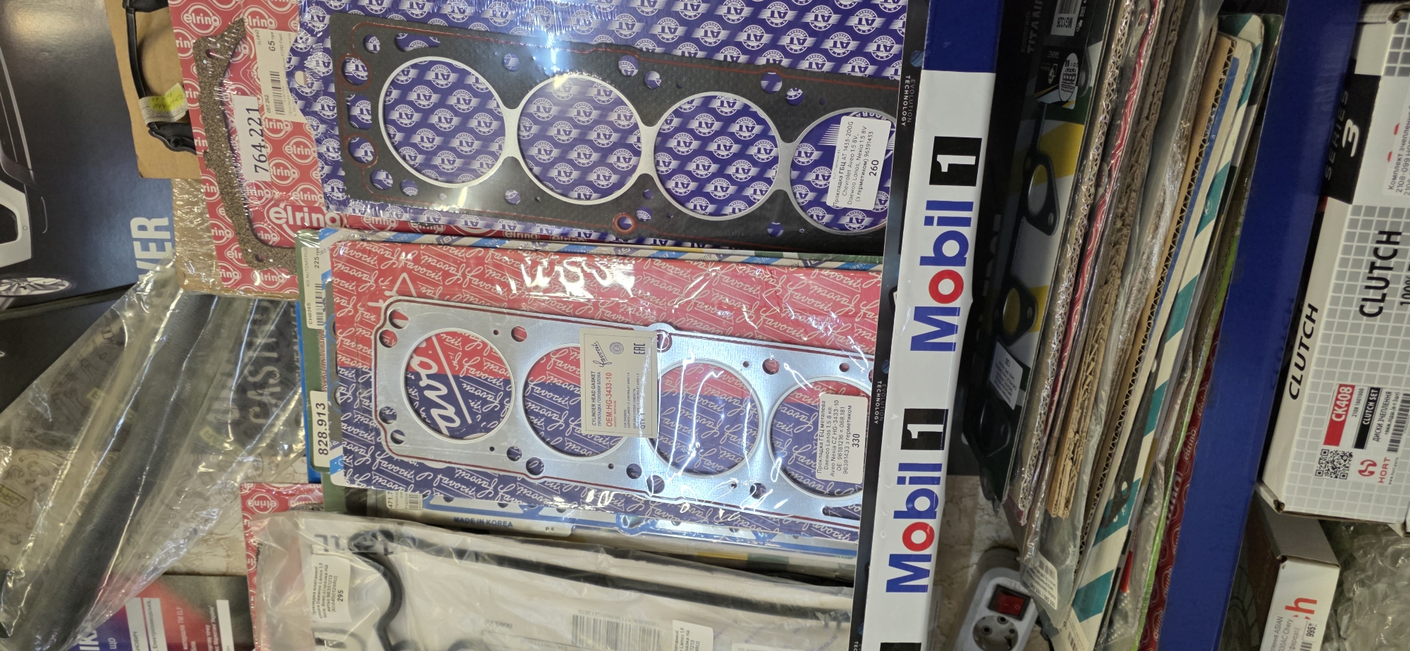 The image shows a shelf in a store, likely an auto parts store. The foreground is dominated by a row of packaged automotive parts, primarily cylinder head gaskets, stacked horizontally. The shelves are blue and white, with the "Mobil 1" logo prominently displayed on the front of the shelf. The background reveals more stacked boxes and plastic-wrapped items, suggesting a cluttered retail environment. The overall impression is of a well-stocked store with a focus on automotive components. The image does not contain any identifiable people or actions. The location is stated to be Kryvyi Rih, Ukraine, but this information is not visually discernible from the image itself.