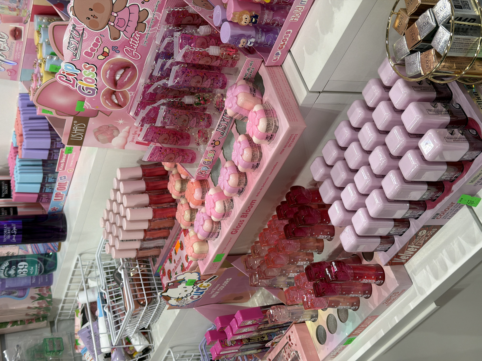This media file captures an indoor retail display of various cosmetics, predominantly lip products and body mists, in what appears to be a store in Izhevsk, Russia. No people are visible.
The scene showcases multiple shelves and display units filled with colorful beauty items. A prominent display in the center features "JSYU Lip Gloss" with a cartoon bear design, labeled "Glitter," along with smaller tubes of glitter lip gloss, some attached to keychains. Below it, another "JSYU" display, "Gloss Bloom," presents flower-shaped lip balms.
To the right, rows of pink, square-bottled lip glosses are stacked, labeled "Velvet Lip Gloss," with a visible price tag of "120-". Below these, clear and reddish lip care products are displayed. Further right and up, small bottles with cartoon figures, labeled "morning" and "Love" lip glosses, also bearing the "Hold me" text, are arranged. A small golden basket contains miniature items, some marked "VMR," likely eyeshadows or pigments.
On the left side, "USHAS" and "Kiss Beauty" branded lip glosses and lip oils, including "Moisturizing" products, are visible. Below them, a section displays various body mists, notably "VICTORIA'S SECRET Love Spell" and other brands like "Nici." The overall aesthetic features pastel colors, glitter, and cartoon characters, suggesting a target audience of younger consumers or those seeking affordable and playful beauty products.