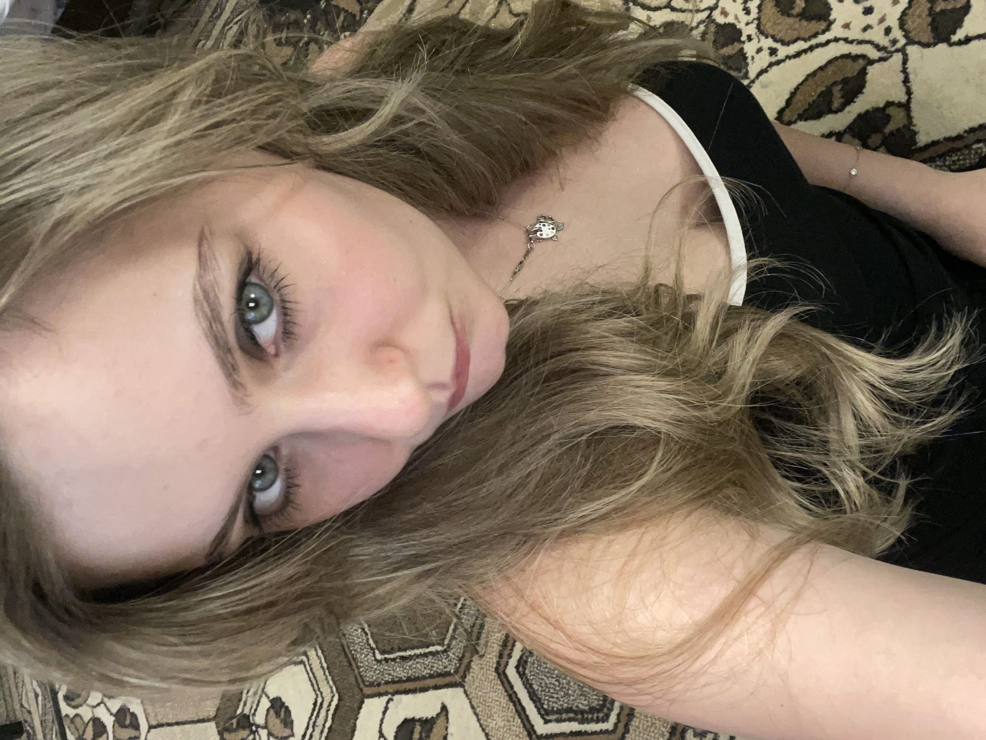 A woman with blonde hair and blue eyes is taking a selfie indoors, possibly on a rug. She is wearing a black tank top with white trim and a silver necklace. The background is out of focus, but appears to be a patterned rug.