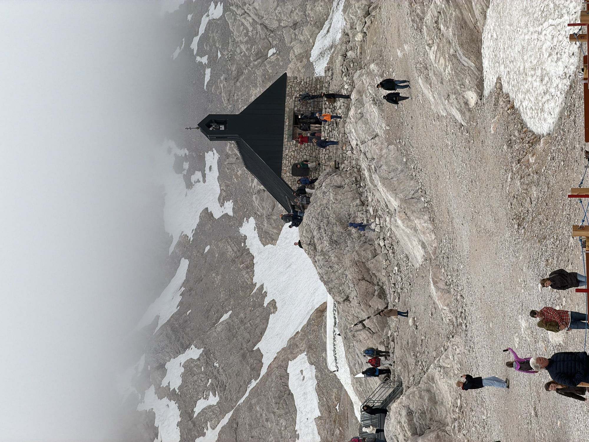 The image depicts a mountainous landscape with a small, dark-colored chapel situated on a rocky outcrop. Snow is visible in patches on the mountain slopes. Several people are scattered around the chapel and on the surrounding rocky terrain, some of whom appear to be tourists. The sky is overcast and foggy, obscuring the upper portions of the mountains. The location is likely a high-altitude tourist area, possibly the Zugspitze, Germany's highest peak, which is near Garmisch-Partenkirchen. The chapel has a stone base and a dark, pitched roof with a small steeple and cross. The ground is covered in gravel and rocks, with some areas of snow. People are seen walking, standing, and taking photographs. One individual is holding a child. The overall atmosphere is cool and rugged, typical of a mountain environment in cloudy weather.