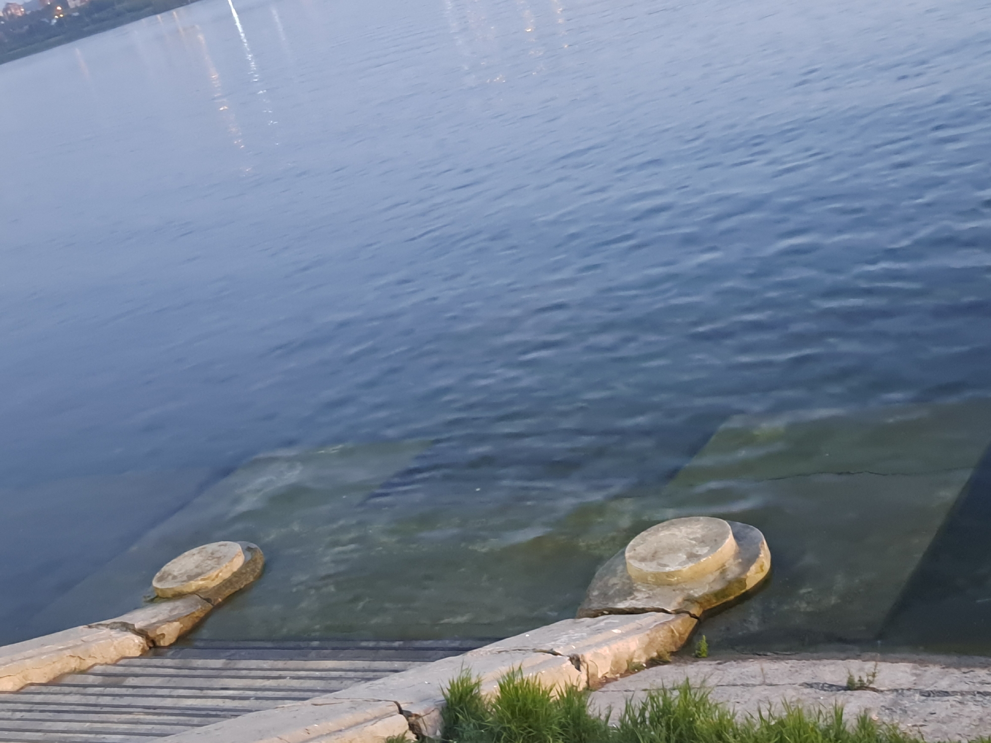 The image depicts a waterfront scene, likely a park or public area, adjacent to a body of water. In the foreground, there are stone steps leading down to the water's edge. Two circular concrete bollards, one partially submerged, are visible along the water line. The water itself appears calm with a slight ripple, reflecting distant lights from what could be buildings or streetlights on the opposite shore. Green vegetation grows along the edge of the steps. The overall impression is one of a tranquil, urban waterside environment. While the image is of a waterfront, it does not provide any specific visual cues to confirm it is located in Irkutsk, Russia.