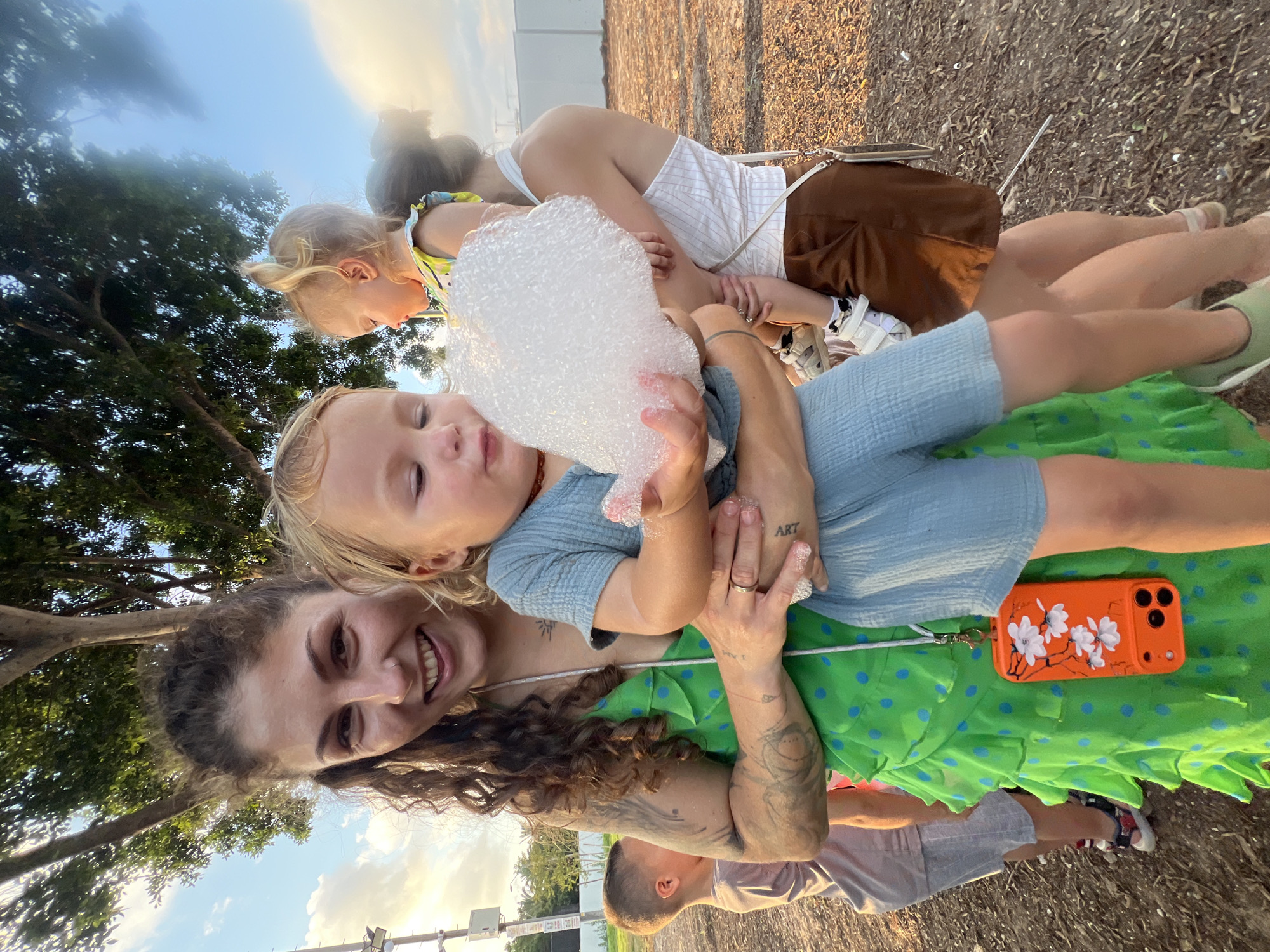 The image shows a group of people, likely a family, outdoors in Kiryat Bialik, Israel. The main subject is a woman holding two young children. One child, a baby, is being held close to the woman's chest, with their face partially obscured by a large bubble. The other child, a toddler with blonde hair, is being held on the woman's lap and is also interacting with the bubbles. The woman is smiling warmly, looking directly at the camera. She has curly brown hair and visible tattoos on her arms and chest. She is wearing a bright green dress with blue polka dots.
A second woman, presumably the mother of the children, is visible in the background, also holding one of the children. She is wearing a white and striped top and brown shorts. The children are dressed in casual summer clothes. The scene appears to be outdoors, with a tree and foliage visible on the left side of the image. The ground is covered in a gravel-like material. The sky is visible in the upper portion of the image, with some clouds and a bright, possibly setting sun. The lighting suggests it is late afternoon or early evening.
A notable object is an orange phone case with a floral design, placed on the woman's green dress. There is also text visible on one of the woman's tattoos, which appears to read "ART". The overall mood of the image is happy and relaxed, with a focus on family interaction and playtime.