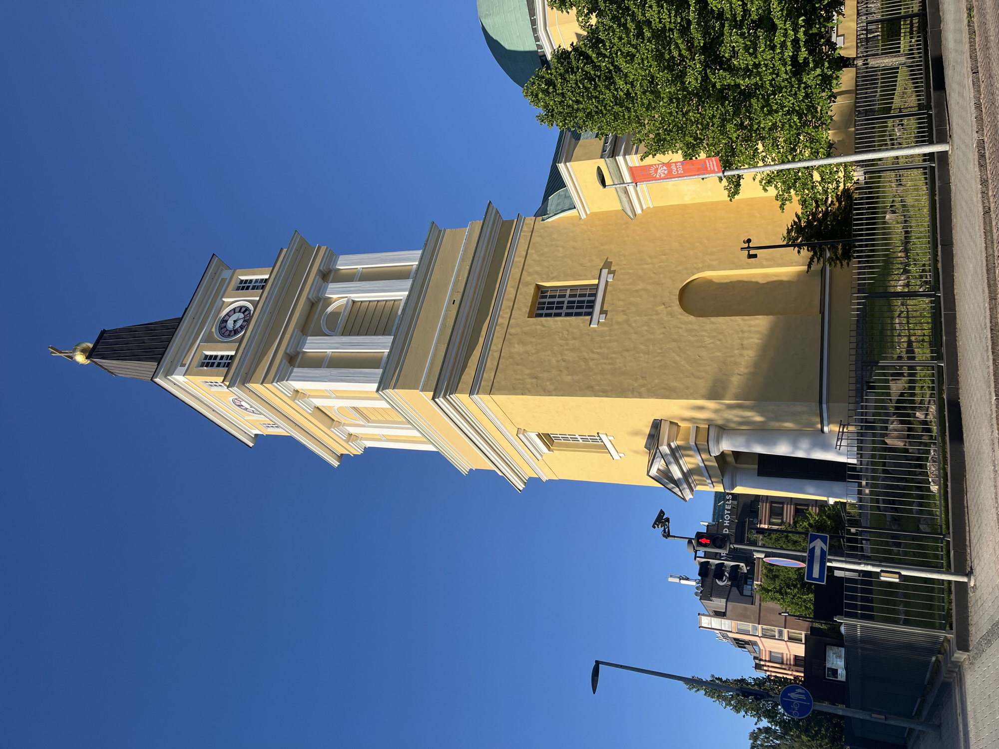 The image shows a sunny day in Oulu, Finland, with a view of the Oulu Cathedral and its clock tower. The cathedral is painted a bright yellow with white trim and has a green dome visible to the right of the tower. The clock tower has a dark spire topped with a golden cross. A large clock face with Roman numerals is visible on the tower. In front of the cathedral, there is a metal fence and some greenery. To the left of the cathedral, there are modern buildings, street lights, traffic signals with a red light illuminated, and street signs. One sign indicates a bicycle path and pedestrian route, and another is a blue arrow pointing right. A red banner with "OULU 2026" is hanging from a pole in front of the cathedral. The sky is clear and blue.