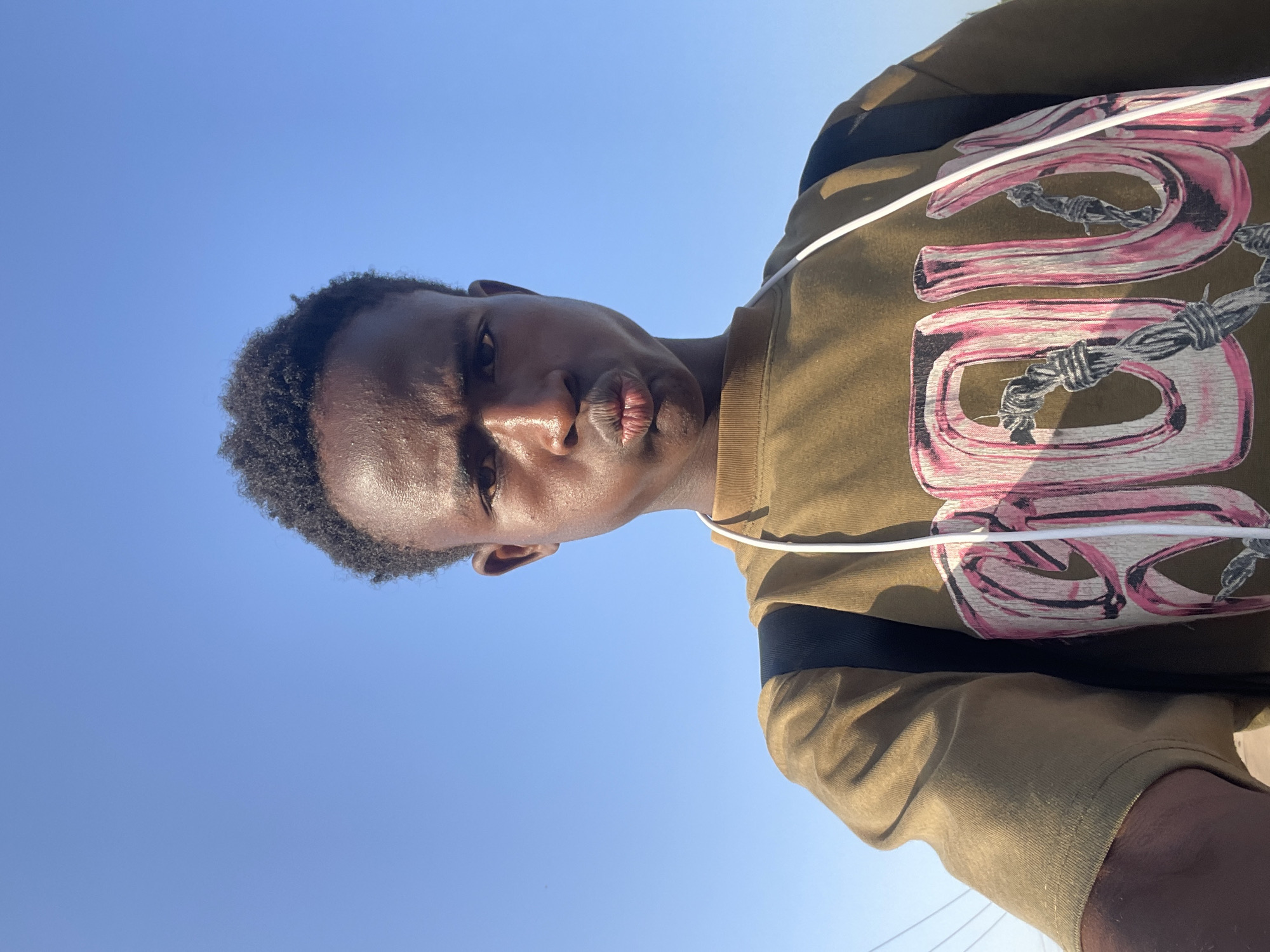A selfie of a young Black male is captured from a low angle against a clear blue sky, suggesting an outdoor setting in Jos, Nigeria. The individual has dark, textured hair styled upwards and is looking directly at the camera with a pouting expression. He is wearing a tan-colored, short-sleeved t-shirt with a graphic print on the front featuring large pink letters interwoven with barbed wire. A black backpack is visible over his shoulders, and a white cord, possibly from headphones, hangs down his chest. The lighting suggests it is daytime, and the background is dominated by the expanse of the sky with some faint power lines visible in the distance on the left side.