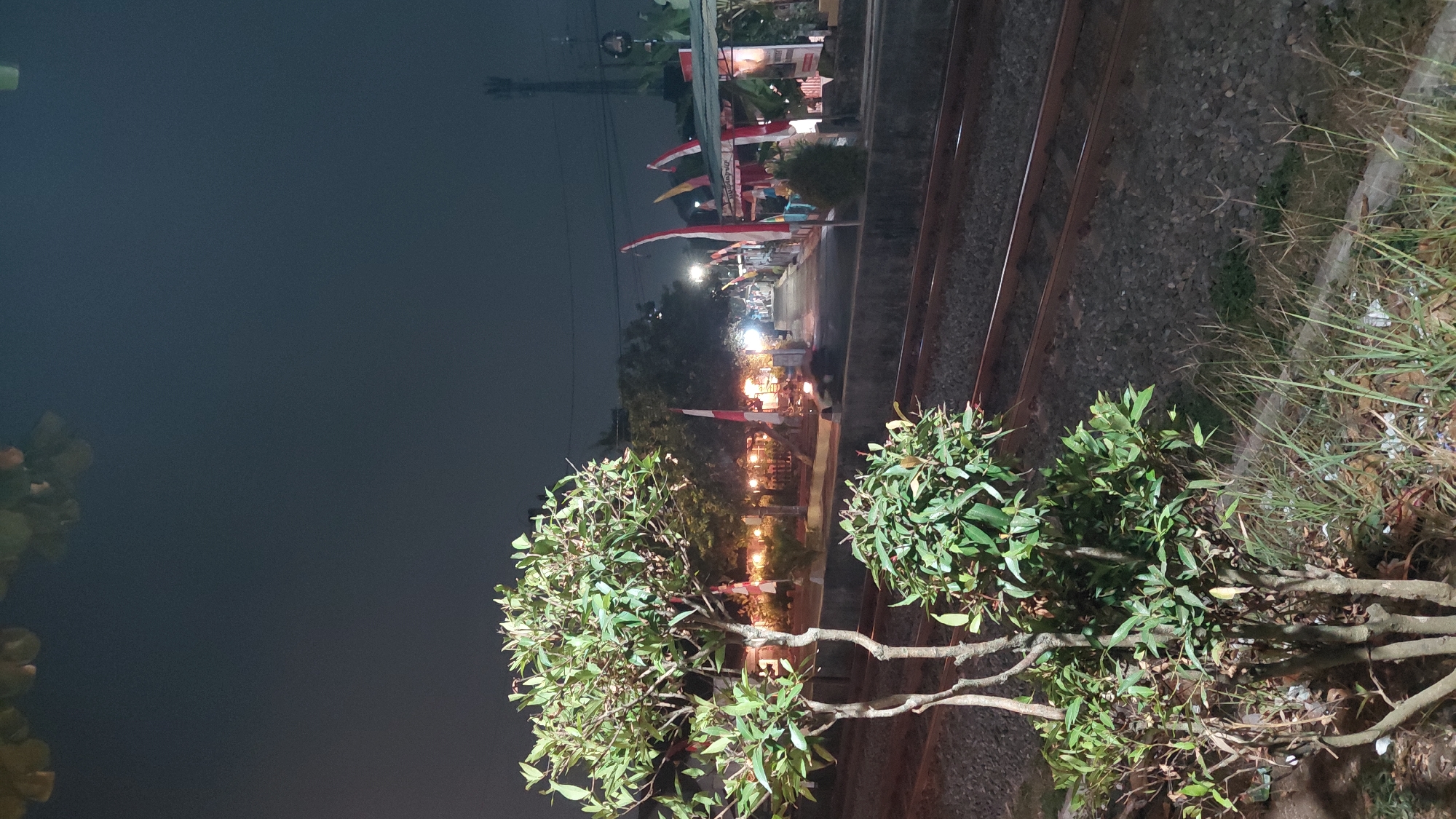 The image depicts a nighttime scene at a railway track in Bantarsoka, Indonesia, likely near a train station. In the foreground, two potted trees with green foliage stand on the left, adjacent to the gravel ballast of the railway tracks. The tracks run horizontally across the lower half of the image. Beyond the tracks, a raised platform or sidewalk lines the railway. Along this platform, numerous Indonesian flags, red and white, are prominently displayed, suggesting a festive or commemorative occasion. Several small structures, possibly food stalls or vendor booths, are visible along the platform, illuminated by warm, yellow lights. In the background, trees form a dark silhouette against the night sky, and a distant cellular tower is visible on the right. A few indistinct figures are present on the platform. The overall atmosphere is dimly lit, with the primary light sources being artificial lights from the stalls and the glow of the flags.