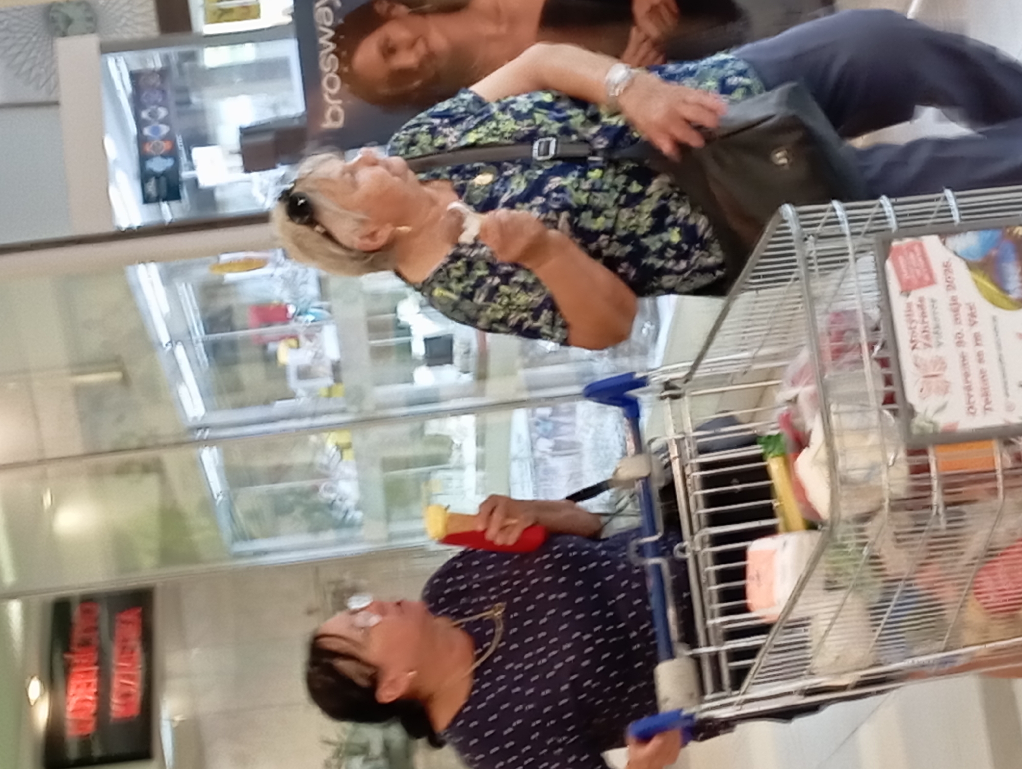 The image shows two women in a shopping cart aisle in what appears to be a supermarket or shopping center. The woman on the left is wearing a dark blue dotted shirt and glasses and is holding a red object with a yellow top, possibly an ice cream cone. She is looking to the right and is pushing a shopping cart. The woman on the right is wearing a floral print shirt and sunglasses pushed up on her head. She is holding a white object in her right hand and has a dark colored purse slung over her shoulder. She is also looking to the right. The shopping cart is filled with various items, including some white and pink packaging. In the background, there are store displays and signage. One sign on the left is red with white text and says "KADERNÍCTVO" and "KOZMETIKA". Another sign on the right has the brand name "brosway". The overall lighting suggests an indoor commercial environment. The image was taken in the city of Nitra, Slovakia.