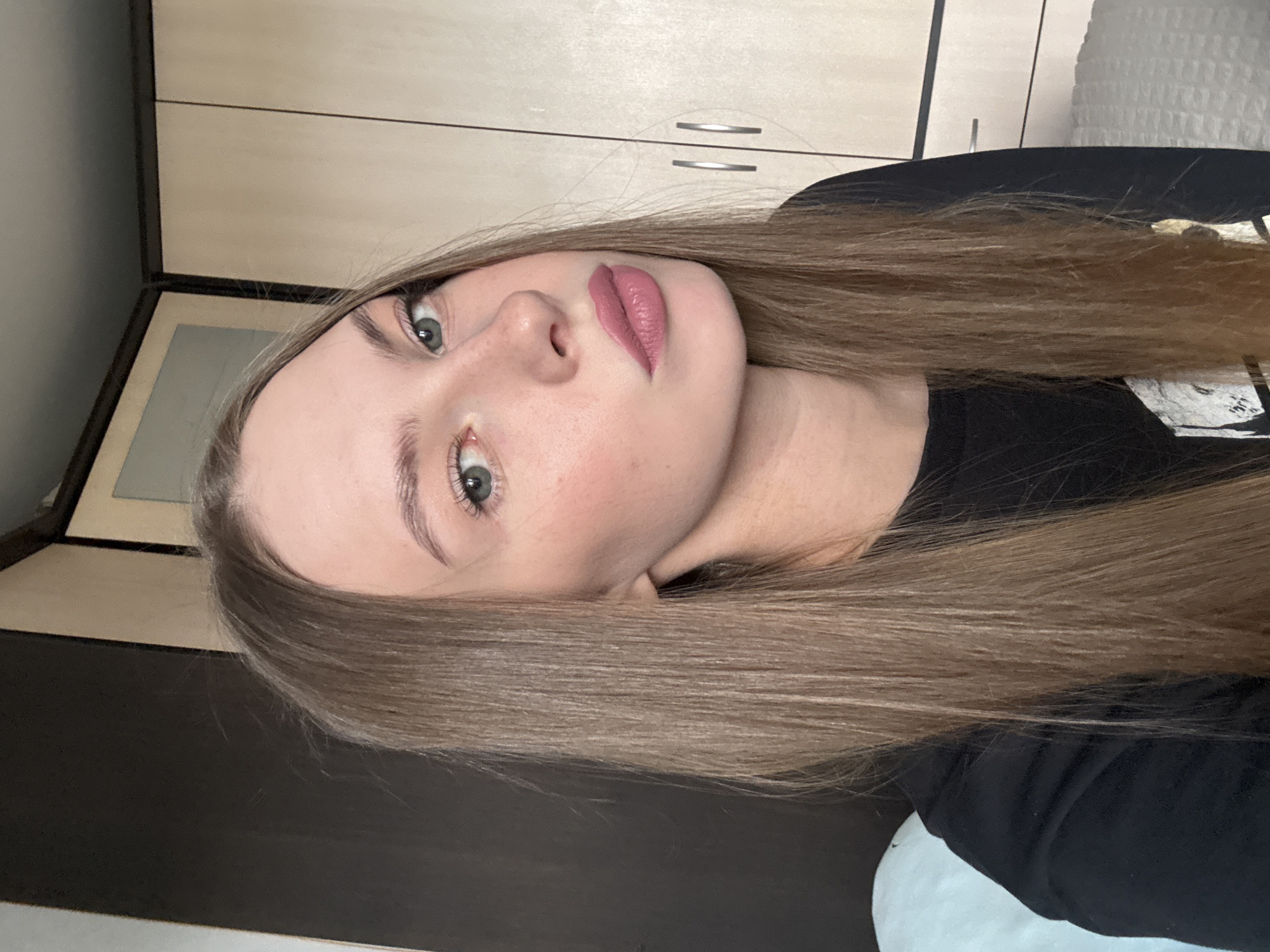 A young woman with long, straight, brown hair and light green eyes is shown from the chest up. She is wearing a black t-shirt. Her lips are painted a matte pink shade. She has blush on her cheeks and her eyebrows are well-defined. She is looking directly at the camera. The background consists of a dark brown wardrobe on the left and a light wood-toned wardrobe with two doors and silver handles on the right. The lighting appears to be natural, possibly from a window. The scene is indoors. There are no discernible landmarks or elements that would indicate a specific location within the city of Perm, Russia.