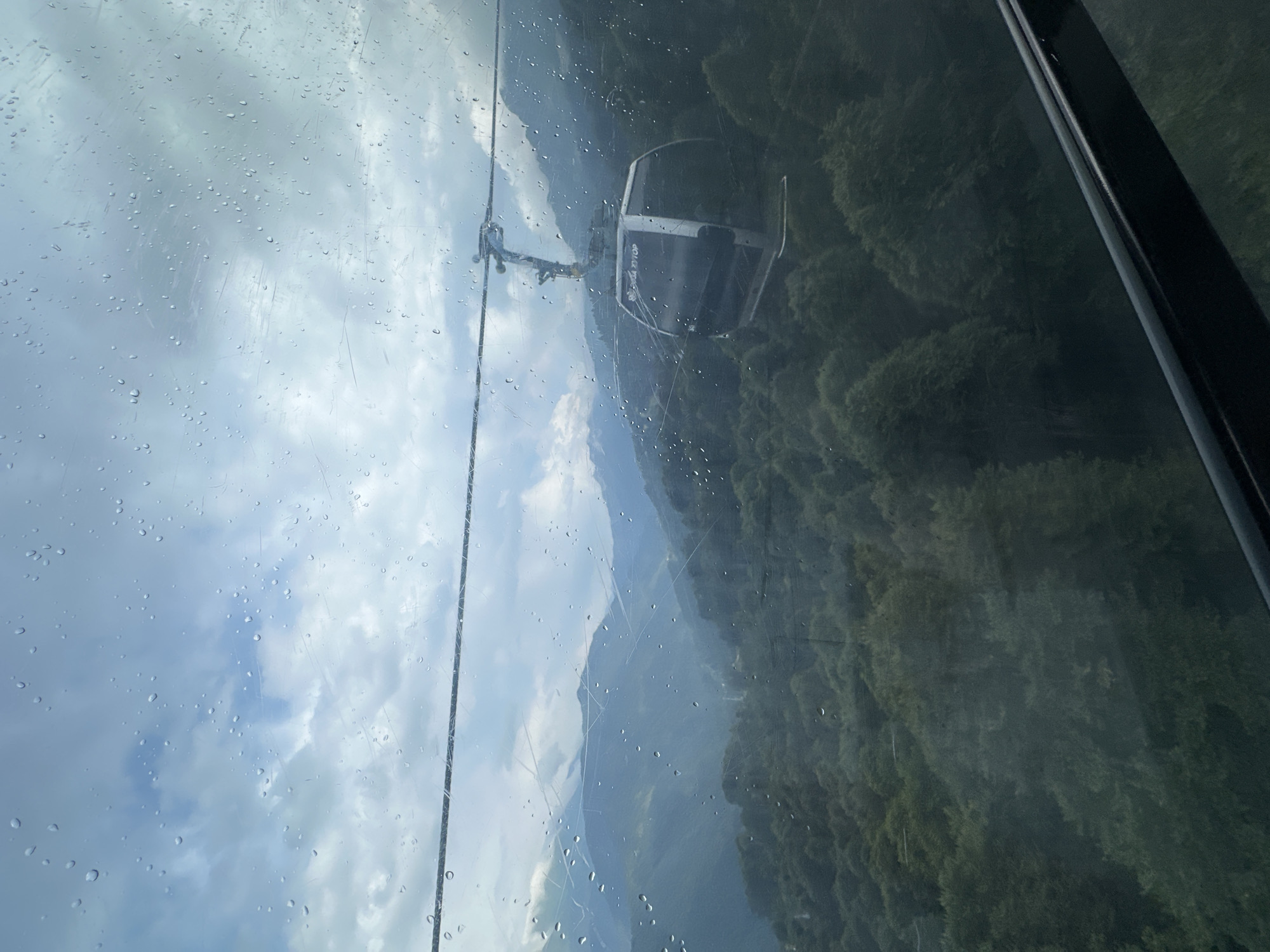 An exterior, slightly elevated shot captures a modern cable car cabin suspended from a wire, traveling through a misty mountain landscape. The cable car is dark gray and white with tinted windows, and the word "ROZA KHUTOR" is visible on its side. It is positioned in the middle ground of the frame. The surrounding environment is dominated by lush, dark green trees blanketing the slopes of rolling mountains. The sky is overcast with patches of blue peeking through the clouds, and a light rain is visible, with numerous water droplets scattered across the foreground, suggesting the photo was taken from inside another moving cable car or enclosed viewing area. The overall scene conveys a sense of travel through a natural, mountainous region, likely a ski resort in Estosadok, Russia, given the prominent signage on the cable car.