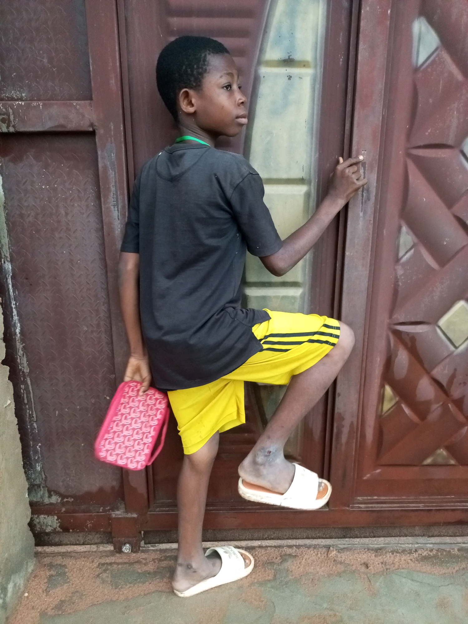 A young Black boy, appearing to be between 8 and 10 years old, stands against a weathered, dark brown metal door. He is positioned with his left leg bent and resting on the doorframe, his right leg extended downwards. He is wearing a black t-shirt with a green collar tag and bright yellow shorts with black stripes. In his left hand, he holds a small, pink purse with a repeating pattern. His right hand is resting on the door near a handle. The door has a diamond-shaped embossed pattern and appears to be old and possibly rusted in some areas. The ground in front of the door is covered with a dusty, sandy material. The scene is outdoors, with natural lighting. The specific location in Gombe, Nigeria, cannot be determined from the image alone.