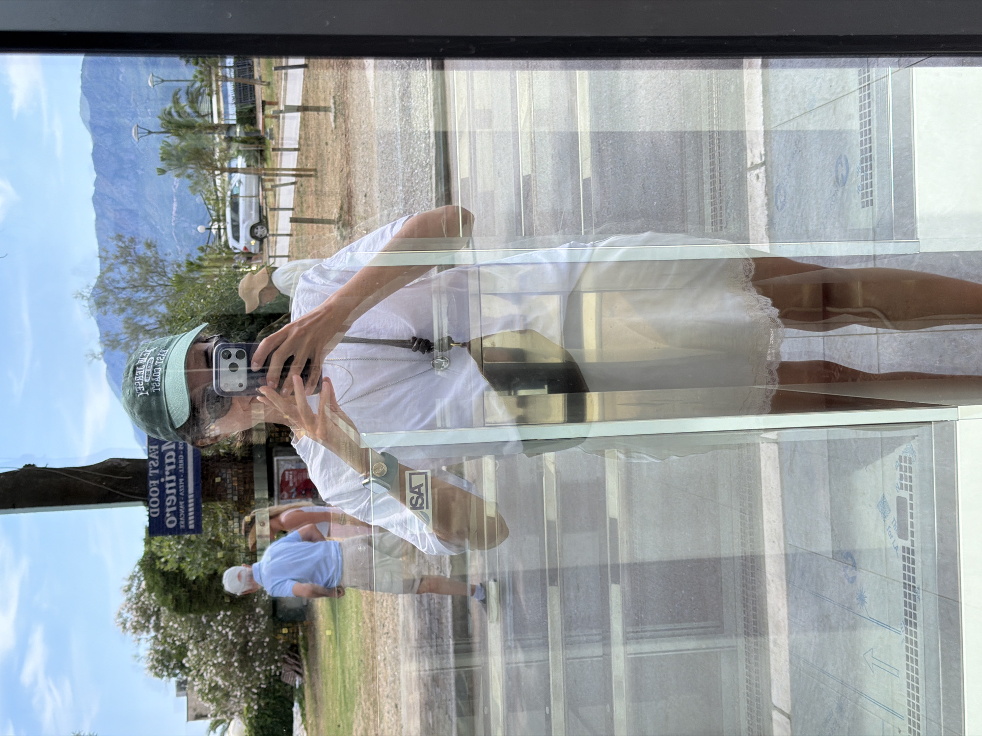 The image is a mirror selfie taken by a woman standing in what appears to be a storefront or cafe with large glass windows. She is wearing a white t-shirt, a white slip dress with lace trim, sunglasses, and a green baseball cap. She is holding a smartphone in her right hand to take the photo.
Reflected in the glass, behind her, is an outdoor scene. The background includes trees, a road, a white SUV, and mountains in the distance. A sign visible in the reflection reads "FAST FOOD MARINERO" and other text in what appears to be Cyrillic or Latin script. This suggests the location is likely in or around Budva, Montenegro, a coastal city known for its historical Old Town and surrounding natural beauty. Two other people are visible in the reflection, one man in a light blue polo shirt and beige shorts, and another person behind him wearing a beige hat and a white top. The ground in front of the storefront has a tiled pattern.