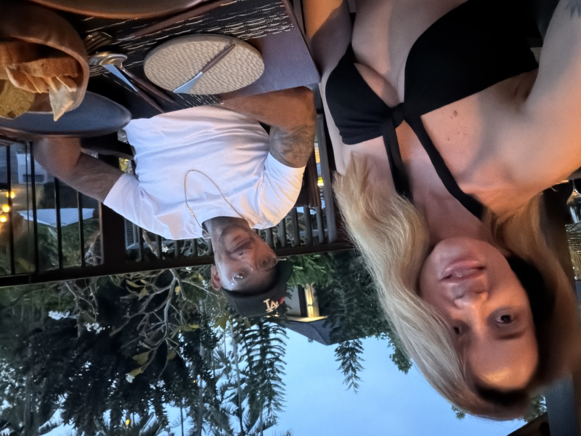 A photograph shows two people sitting at a table set for a meal, presumably at an outdoor restaurant in Kediri, Indonesia, based on the tropical foliage and architecture visible in the background.
The man, on the left, wears a white t-shirt, a gold chain, and a baseball cap with the "LA" logo. The woman, on the right, has blonde hair and wears a black top.
The table features a placemat, plate, knife, and a wooden bowl containing bread or chips. Beyond the table, a metal railing is visible. The sky in the background suggests it is dusk or early evening.