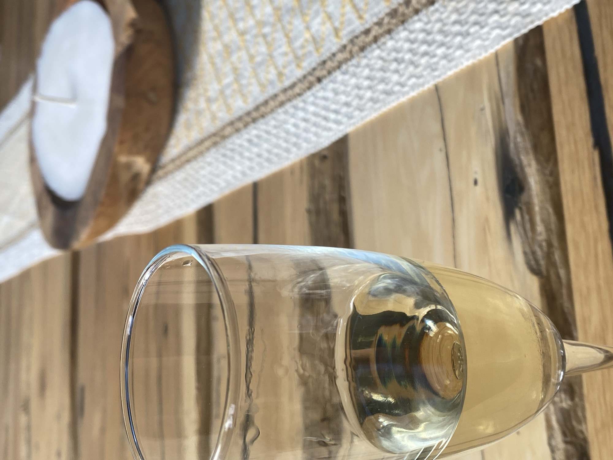 A close-up shot focuses on a glass of white wine on a wooden table. To the right, a candle in a wooden holder rests on a placemat with a yellow and white pattern. The background is blurred, suggesting an outdoor setting in Krasnoflotskoe, Russia. The lighting indicates it is daytime.