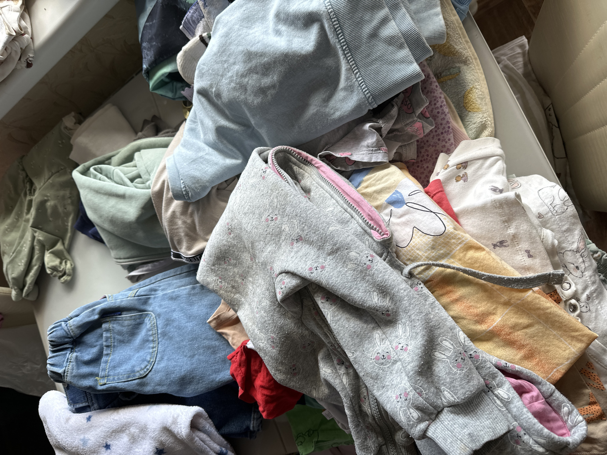 The image shows a pile of folded and unfolded children's clothing, including shirts, pants, and a zippered hoodie, situated inside a white storage container or shelf. The clothing is a mix of colors and patterns, including a gray hoodie with a pink zipper lining, decorated with bunny motifs. The location is likely indoors, and no information in the image suggests the city of Nizhny Novgorod, Russia.