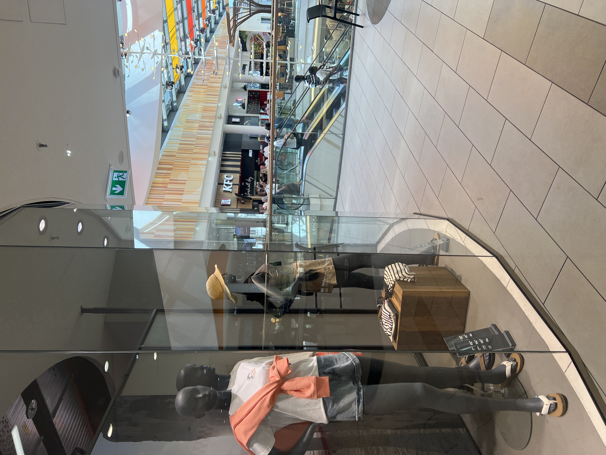 The image depicts the interior of a modern shopping mall, likely in Aachen, Germany, given the presence of a KFC outlet visible in the background. The scene is captured through a glass display window, showcasing mannequins dressed in casual attire.
In the foreground, two mannequins are displayed. The left mannequin is wearing a white t-shirt, denim shorts, and has an orange sweater tied around its shoulders. The right mannequin is wearing a striped top and brown shorts, with a straw hat. Below the mannequins, a wooden display box holds folded clothes. A price list is also visible in front of the mannequins.
In the background, several people are using escalators. Various retail outlets, including a KFC restaurant with signage indicating "Fresh all day", are visible. The mall features a contemporary architectural style with a high ceiling, modern lighting fixtures, and decorative elements on the walls. The floor is tiled. The overall atmosphere suggests a typical bustling shopping environment.