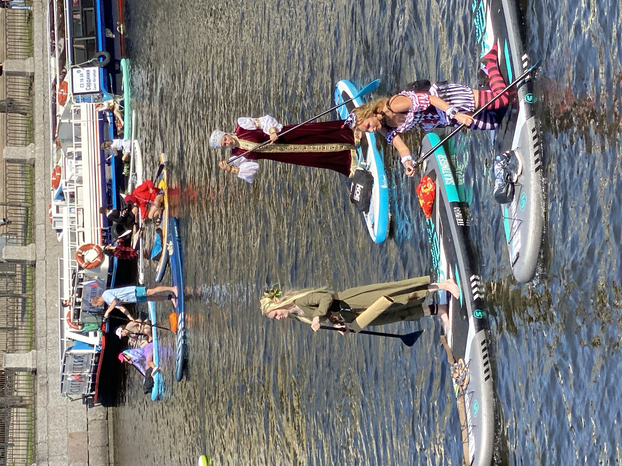 The image shows a group of people in costumes on paddleboards in a canal in Saint Petersburg, Russia. The canal is lined with a stone embankment and a metal fence. In the background, there are several tour boats docked. The paddleboarders are dressed in a variety of historical and fantasy-themed costumes. One woman is dressed in a long, medieval-style gown with a headdress. Another woman is dressed in a jester-like costume with striped stockings. A man in the background is wearing a red hooded costume. The water is dark and appears to be slightly choppy. The sky is bright and sunny.