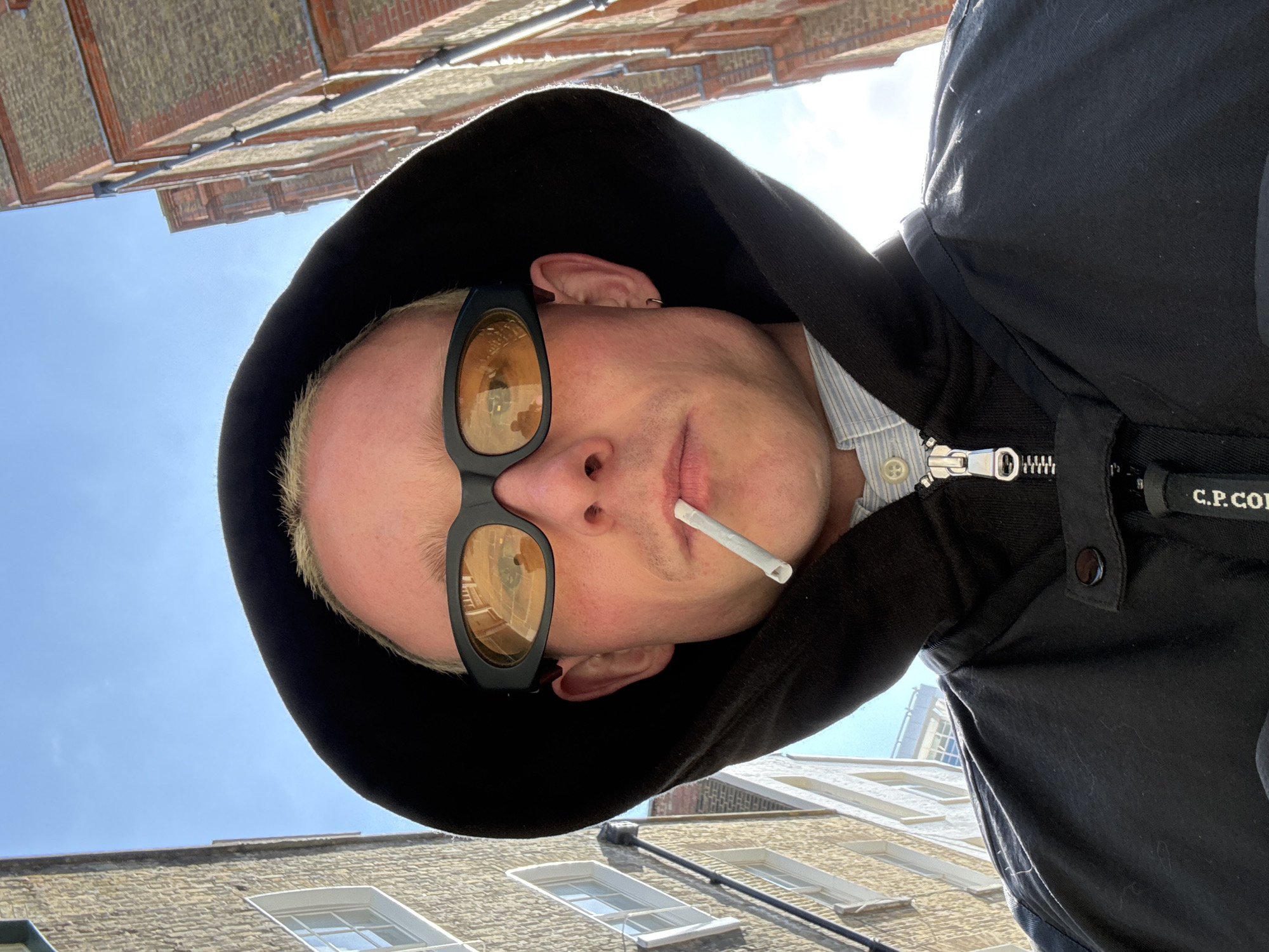 An extreme low-angle, upward-facing selfie captures a person against a bright blue sky and historic buildings, likely in London. The subject, appearing to be an adult with short blonde or light-colored hair and pale skin, is wearing a black hooded jacket. The hood frames their head, and the jacket features a prominent silver zipper and a tag visible on the right side with partially legible text, appearing to say "C.P. CO".
They are wearing distinctive black, wrap-around sunglasses with amber-tinted lenses, which reflect the surrounding architecture and sky. A small, white cigarette is held between their lips. The person has a neutral expression, with light stubble visible on their chin and upper lip. A small silver ear piercing is visible on their right ear.
The background consists of traditional brick and stone buildings, characteristic of London architecture, with several windows visible. The clear blue sky indicates a sunny day. The unique perspective makes the buildings appear to loom overhead, creating an immersive, worm's-eye view. The bright sunlight creates strong contrasts, highlighting the subject and the upper parts of the surrounding structures.