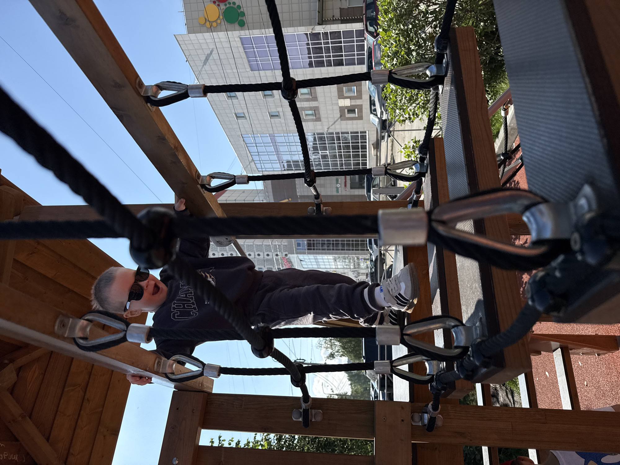 A young boy wearing sunglasses and a dark grey sweatshirt and pants is walking on a wooden playground structure with rope nets. He is smiling with his mouth open. The playground is outdoors in a city. In the background, there is a modern, multi-story building with a glass facade and a blue sky. Several cars are parked in front of the building. This scene is located in the city of Perm, Russia.
