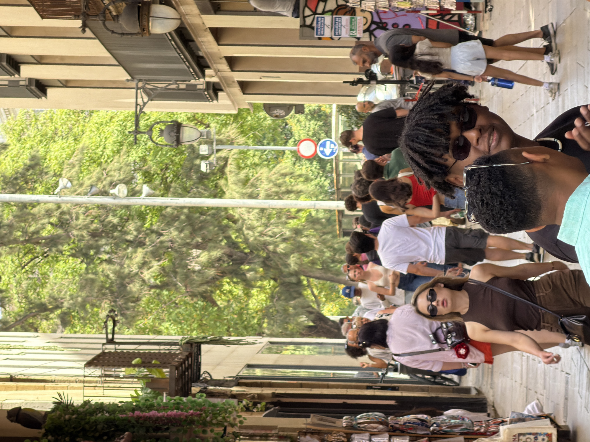 This media file depicts a bustling street scene in Barcelona, Spain. A diverse group of people are walking and interacting on a sidewalk lined with buildings and trees. The sunlight suggests it is daytime, possibly late morning or early afternoon, and the clear sky implies fair weather.
The subjects include individuals of various ages, ethnicities, and styles, many of whom appear to be tourists or locals enjoying a day out. They are dressed casually, with some wearing t-shirts, shorts, and sunglasses, indicative of warm weather. Among the prominent figures is a young woman with dark, braided hair and sunglasses, looking towards the camera with a relaxed expression. Another woman, wearing a brown top and sunglasses, is looking upwards, with a handbag slung across her body. A man with a grey beard and a grey t-shirt is also visible, as are many other individuals in the background, forming a lively crowd.
Objects in the scene include streetlights, traffic signs (a circular sign with a red border and a white center, and another circular sign with a blue center and a white downward arrow), and what appear to be market stalls or shop displays further down the street. The buildings are multi-story structures with balconies and awnings. Lush green trees provide shade and a natural element to the urban environment.
The activity appears to be casual strolling and shopping, with people moving in various directions along the pedestrian walkway. The overall atmosphere is one of everyday life in a vibrant city. The presence of Spanish language signs on some of the stalls or buildings would further confirm the location, though specific text is not clearly legible. The scene is alive with movement and the casual interactions of people enjoying their surroundings.