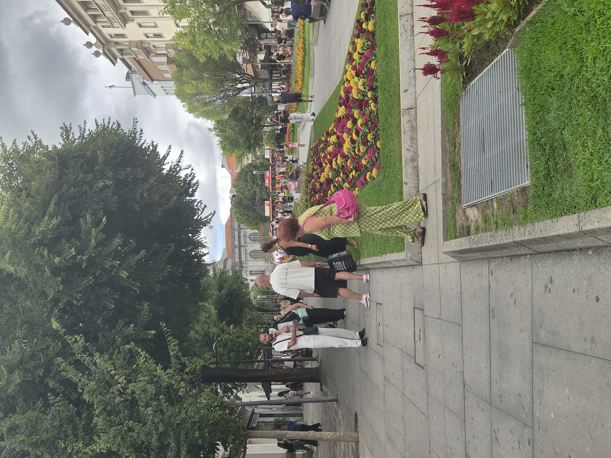 A street scene in Braga, Portugal, features a pedestrian walkway lined with trees. Several people are visible on the sidewalk and in the distance. In the foreground, a woman wearing a green patterned outfit and carrying a pink handbag stands near a flowerbed filled with red and yellow flowers. A man in a white t-shirt and shorts is talking on his phone, while another man in a white t-shirt and white pants stands nearby. In the background, buildings with balconies and ornate architecture are visible, along with more trees and a cloudy sky. The image captures a typical urban environment with people engaged in various activities.