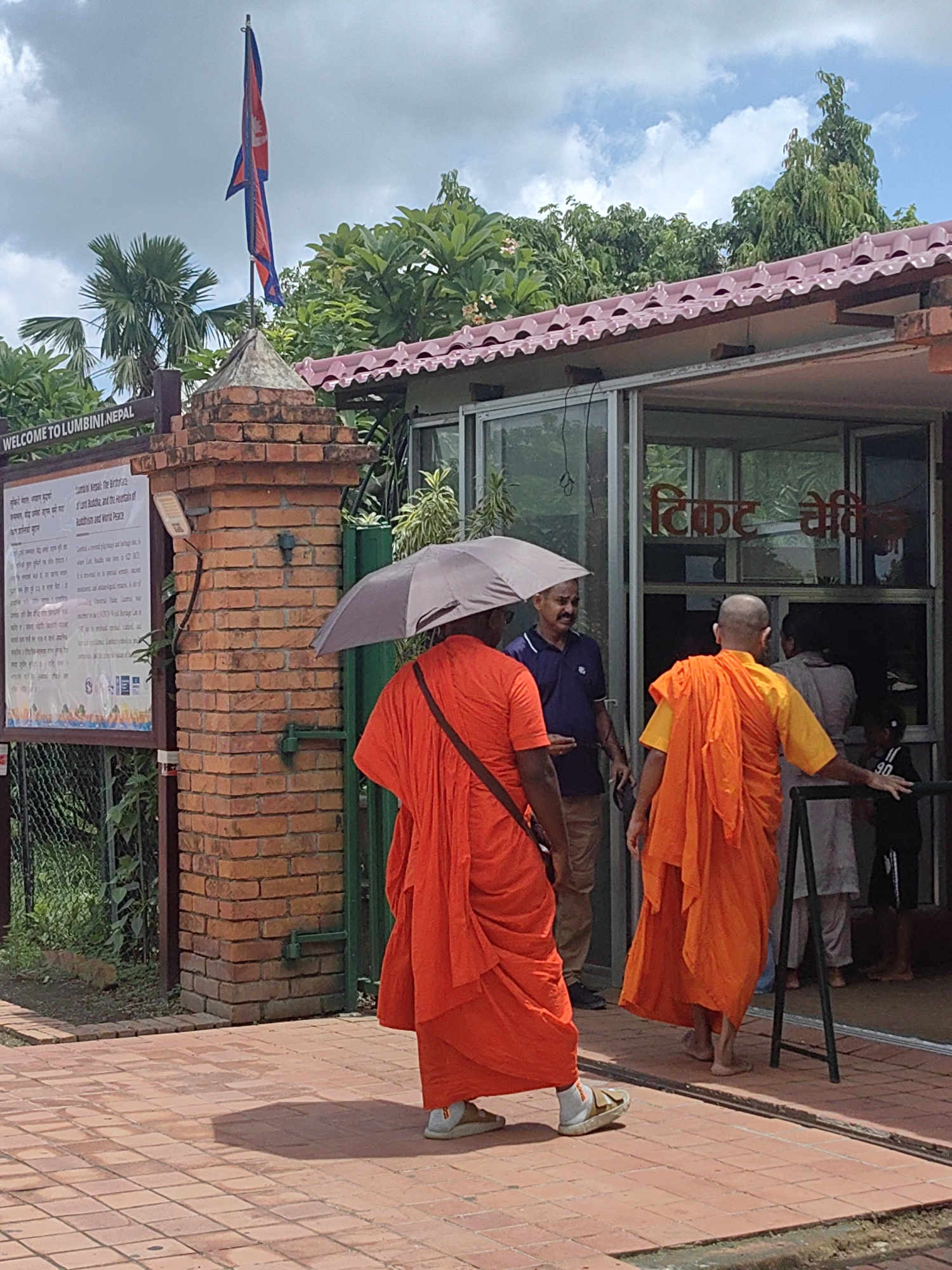 The image depicts an outdoor scene at the Lumbini Bazaar in Nepal. In the foreground, two individuals in bright orange robes are visible. One, on the left, is holding a light-colored umbrella and walking away from the camera. The other, on the right, is facing away from the camera and appears to be a monk with a shaved head, wearing similar orange robes over a yellow undergarment. He is barefoot.
Behind them, a glass-fronted building with a pink tiled roof is present. Red lettering is visible on the glass, possibly indicating a ticket booth or entrance. Several other people are inside or near the building, including a man in a blue shirt and beige pants, and partially visible figures in the background. A brick pillar with a sign board in English and another language stands to the left of the entrance. The sign board has the text "WELCOME TO LUMBINI, NEPAL". A Nepalese flag is flying from a flagpole atop the brick pillar. The ground is paved with reddish-brown tiles. Lush green trees and foliage surround the building and the entrance area. The sky is partly cloudy.