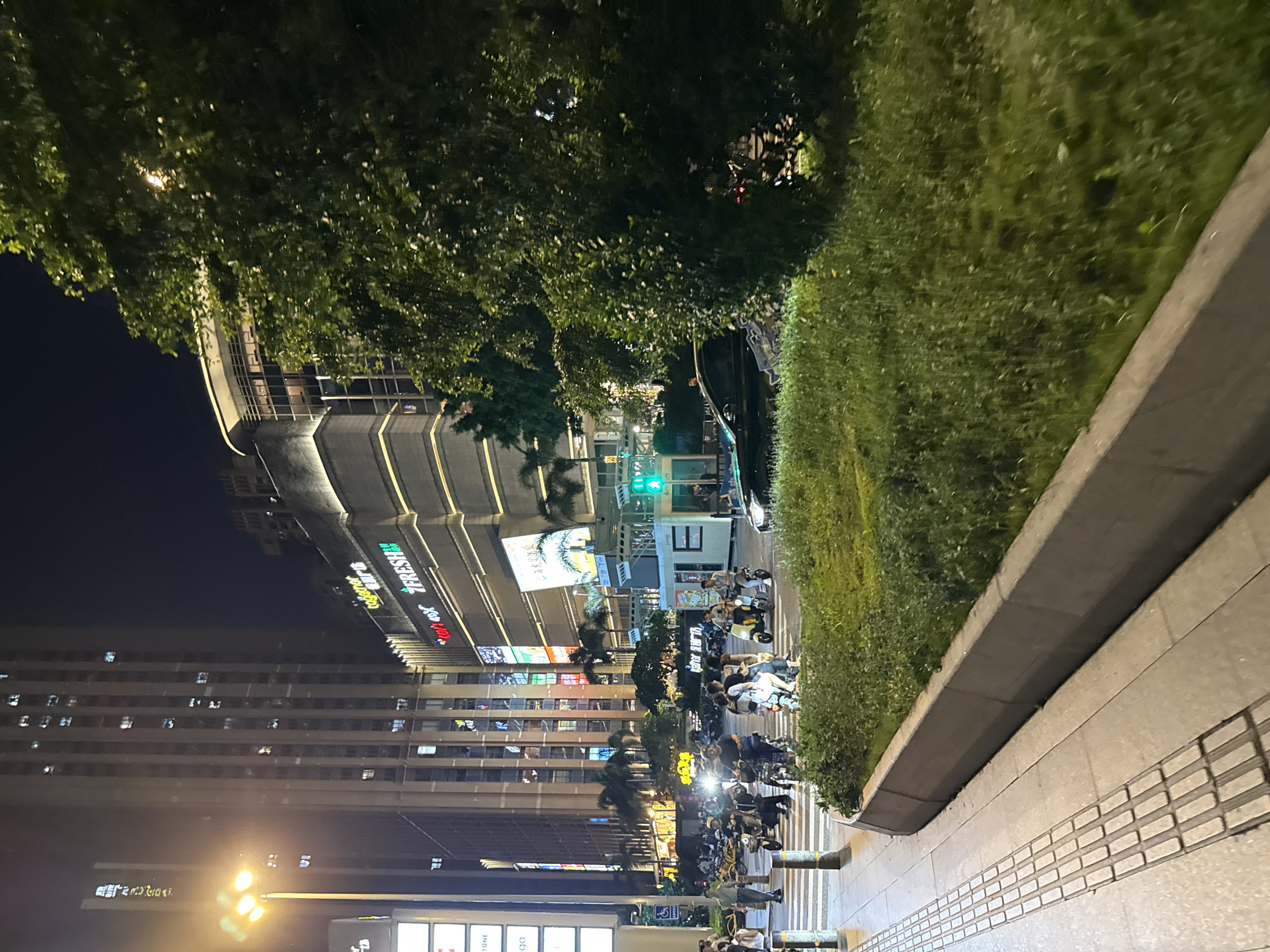 This is a nighttime exterior shot of a busy city street in Haizhu District, China. The scene depicts a pedestrian crosswalk with several people on electric scooters or motorcycles waiting or moving through. Tall buildings with illuminated windows line the background, indicating a commercial or residential area. A large, leafy green tree is prominent on the right side of the frame, partially obscuring a dark-colored sedan. The streetlights cast a yellow glow, illuminating the sidewalk and crosswalk. The overall atmosphere is that of urban nighttime activity.