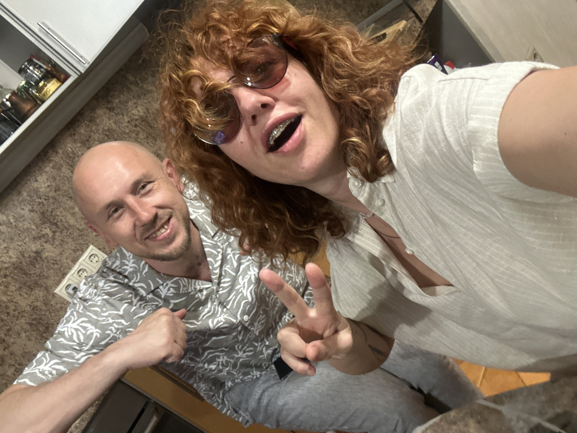 The image depicts a selfie-style photograph of two individuals, one female and one male, captured indoors within what appears to be a kitchen or dining area. The location in Sochi, Russia, cannot be confirmed from the visual evidence present in the image.
The female subject is positioned in the foreground on the right. She has reddish-brown curly hair and is wearing large, dark-tinted round sunglasses. Her mouth is open, revealing dental braces on her upper teeth, and she exhibits a slight smile. She is wearing a light-colored, possibly off-white or light grey, collared shirt with vertical stripes, unbuttoned at the neckline. A subtle necklace, possibly with a single pearl, is visible at her throat. Her left hand is raised, with the index and middle fingers extended in a "peace" sign.
The male subject is standing behind and to the left of the female. He has a bald head and a light stubble beard, and is smiling broadly. He is wearing a grey short-sleeved collared shirt with a white tropical leaf pattern and light-colored, possibly light grey, striped trousers. His right arm is bent at the elbow, with his fist clenched and slightly raised, suggesting a playful flex or pose.
The background elements indicate an indoor setting. To the left of the male's head, a triple European-style power outlet (Type F) is mounted on a light-colored wall. Above and to the right of the male, a white cabinet with a silver handle is partially visible. Inside the cabinet, several jars and containers can be observed, one with partial Cyrillic text visible, reading "MYC". The overall lighting is bright and even, casting soft shadows.