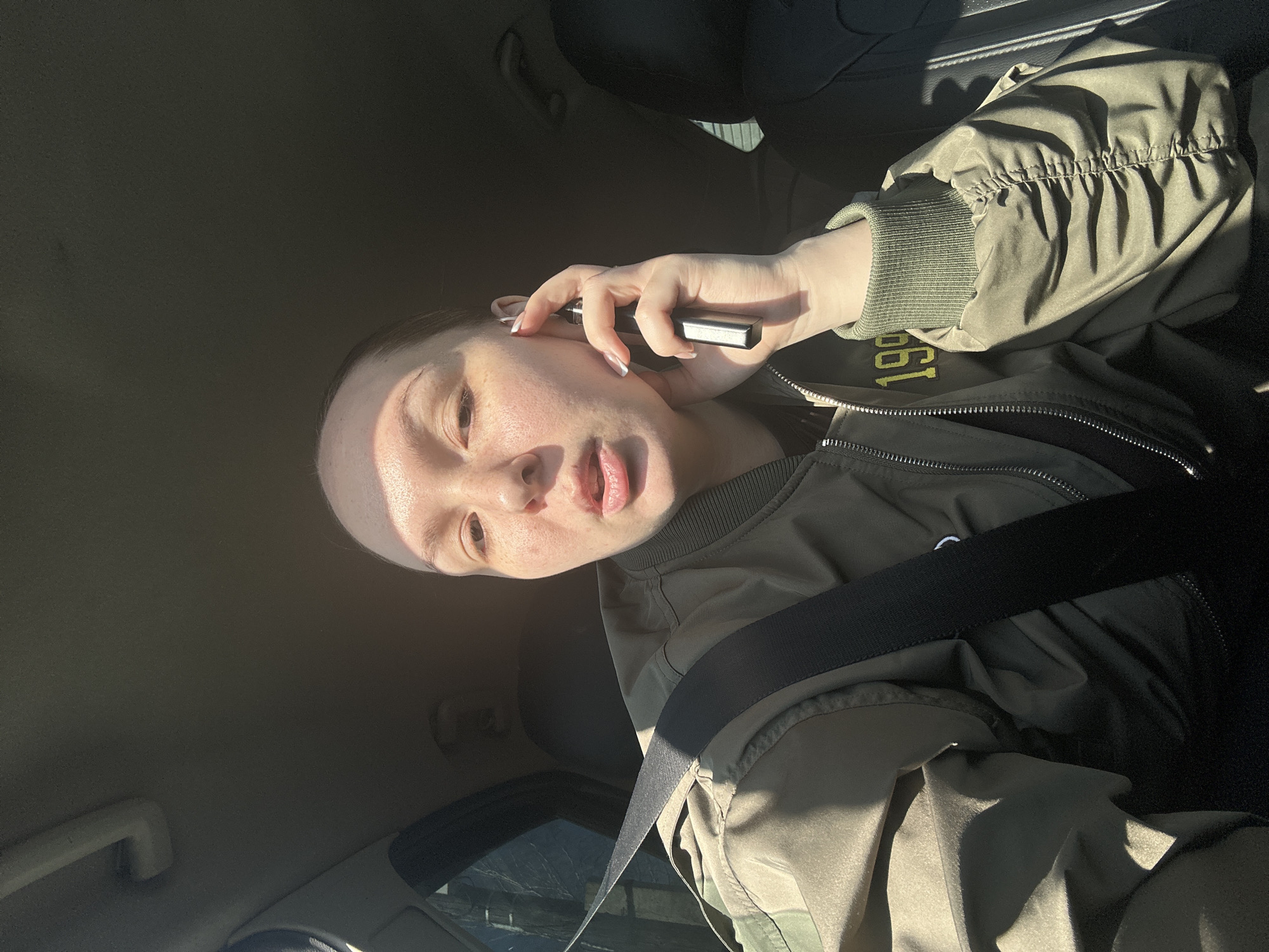 A light-skinned person is shown in a selfie-style photograph taken inside a car. The person is wearing a bomber jacket with the number "199_" embroidered on the sleeve and a seatbelt across their chest. They are holding a small, dark, rectangular object, possibly a vape pen, near their ear. The background shows the interior of the car. The location is not discernible from the visual information provided.