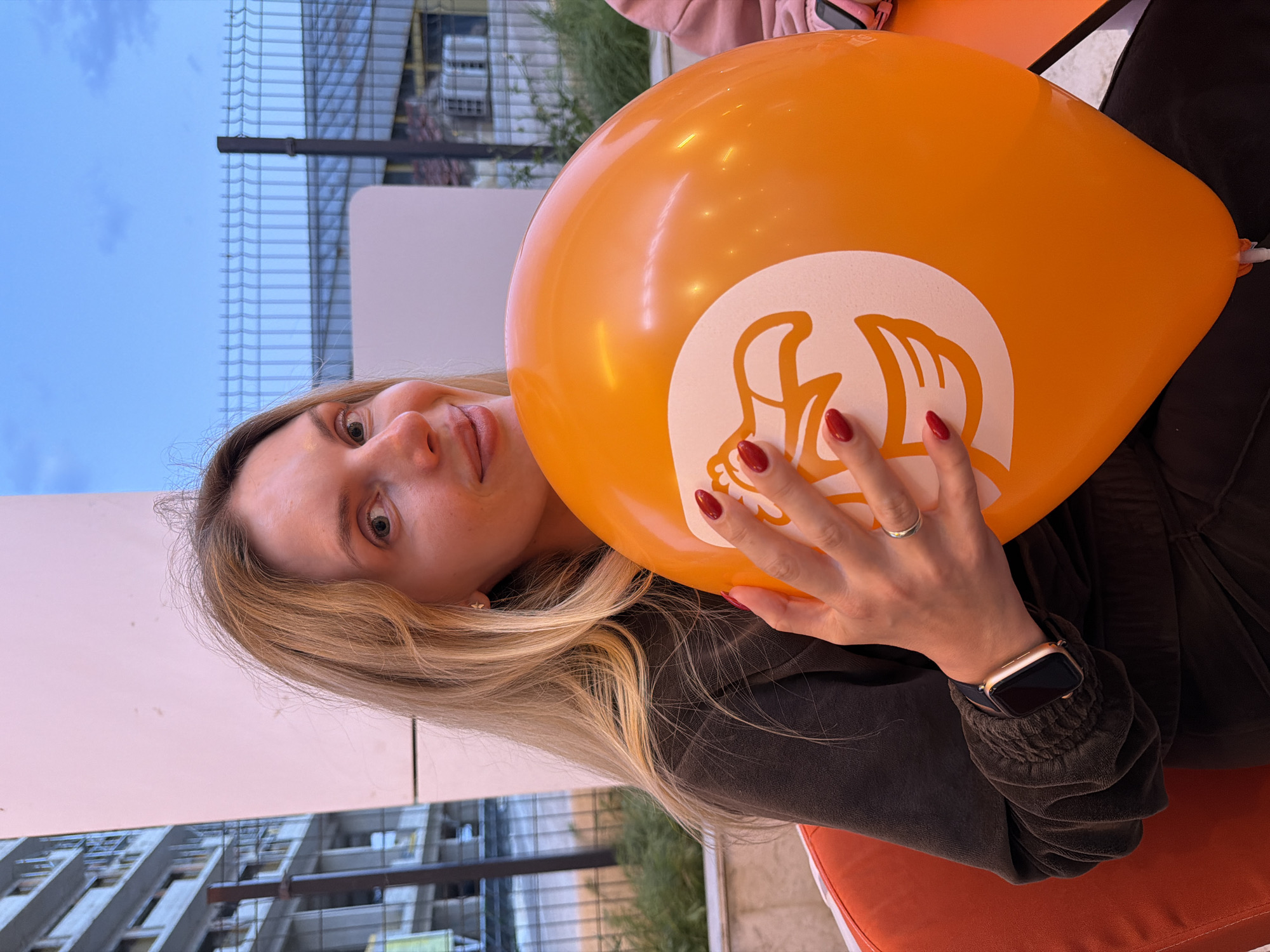 A blonde woman is holding an orange balloon with a white logo in front of her face. She has a watch on her left wrist and a ring on her ring finger. The balloon is obscuring part of her face. The background shows a building, a fence, and the sky.
Based on the architecture and the fence style in the background, this photo was likely taken in or near Budva, Montenegro, specifically in an area with modern construction or renovated buildings.