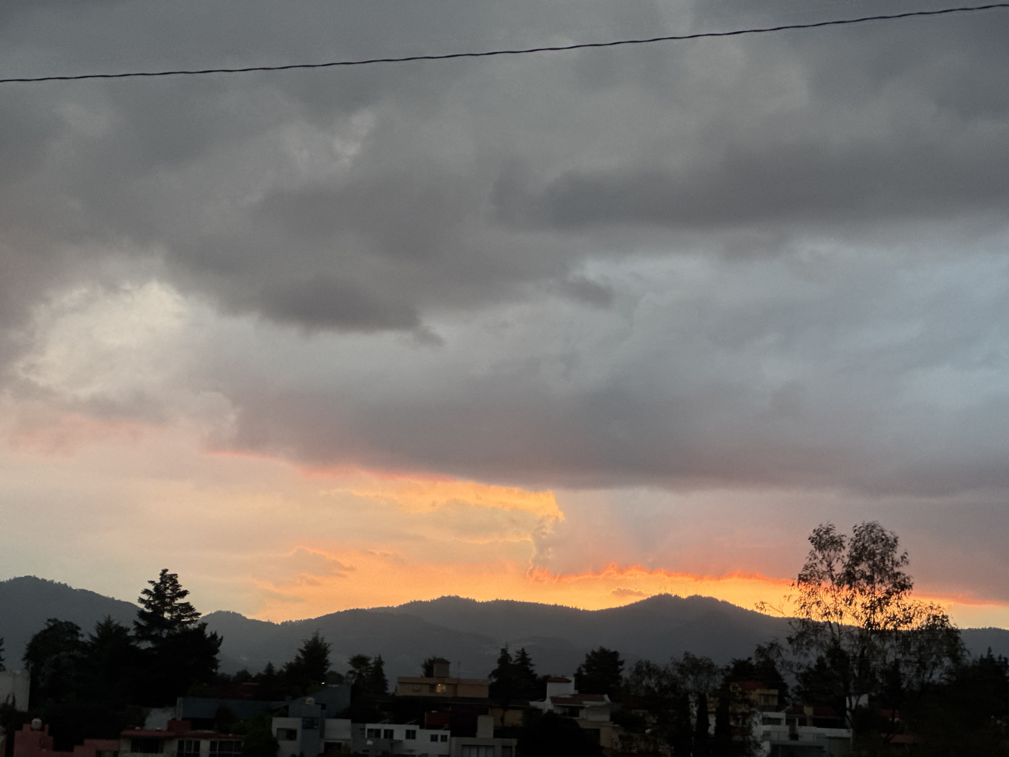 The image depicts a dusky scene in Mexico City, Mexico. The sky is overcast with grey clouds dominating the upper portion of the image. There's a streak of orange and pink light visible beneath the clouds, likely from the setting sun. In the mid-ground, the silhouette of a mountain range is discernible. The foreground features houses and buildings nestled among trees. A power line bisects the top of the image. There are no people visible in the scene.