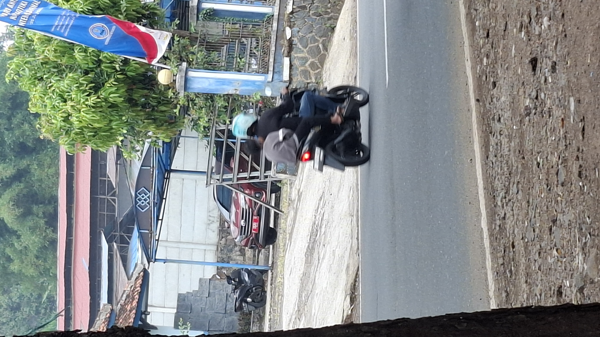 The image captures a street scene in Cilaku, Indonesia, featuring two individuals riding a motorcycle on a paved road. The motorcycle is black and is in motion, with the rear brake light illuminated. The rider wears a dark jacket and blue jeans, and the passenger is seated behind them wearing a gray shirt. The passenger has a light blue helmet on. In the background, a red car with a license plate that appears to read "B 1070" is parked behind a metal fence. Further back, there are buildings with red roofs, a blue canopy structure, and a blue and white flag with red accents waving. Lush green trees and vegetation are visible on a slope behind the structures. The ground beside the road is rough and appears to be covered in stones and debris.
