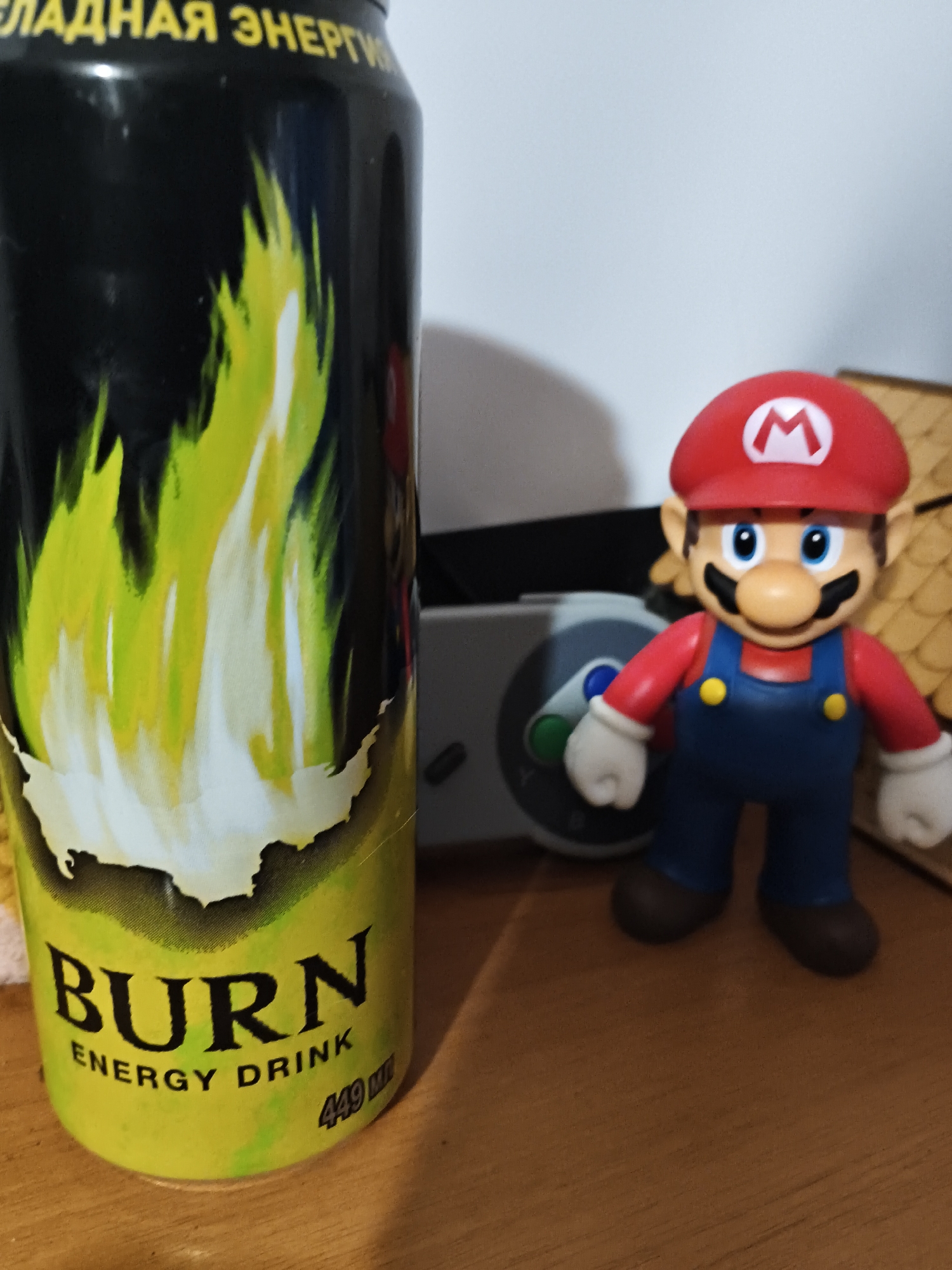 An energy drink can with a green and black design featuring flames and the word "BURN" is positioned on the left side of the frame. To the right of the can is a figurine of the video game character Mario, standing on a wooden surface. The background is indistinct, appearing as a wall. The scene is indoors, on a wooden table. There is no indication of the location being in the city of Mokshan, Russia.