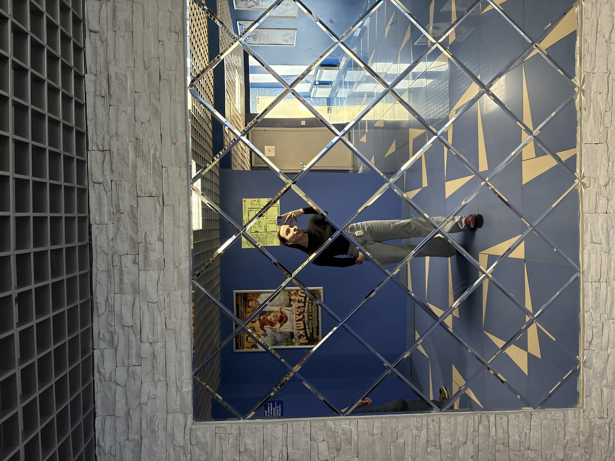 An interior scene in Votkinsk, Russia, features a person taking a selfie in a large, decorative mirror. The mirror, composed of beveled square tiles arranged in a diamond pattern, occupies the right two-thirds of the frame. To the left, a textured wall of light grey, rough-cut stone panels adjoins a dark grey wall or shelving unit with a grid of square compartments.
The mirror reflects the opposite side of the room. A person, dressed in a black long-sleeved top, light grey jeans, and red sneakers, holds a smartphone to capture their reflection. They are looking into the phone, focused on the selfie. Behind them, a vibrant royal blue wall is visible. On this wall, a prominent framed poster features cartoon-style characters, suggesting a movie or theatrical performance. Inverted Cyrillic text is visible on the poster, with some characters hinting at "ЧЕЛОВЕК-ПАУК" (Spider-Man), indicating the location might be a cinema or entertainment venue. Below the main poster, a smaller framed item, possibly a floor plan or informational sign, also displays inverted Cyrillic text.
Above the blue wall, sections of a ceiling with rectangular light fixtures are reflected. The reflected floor is light-colored, possibly beige or light yellow, with a repeating geometric pattern in a darker blue or grey. The bright, even lighting suggests an indoor setting with strong artificial illumination.